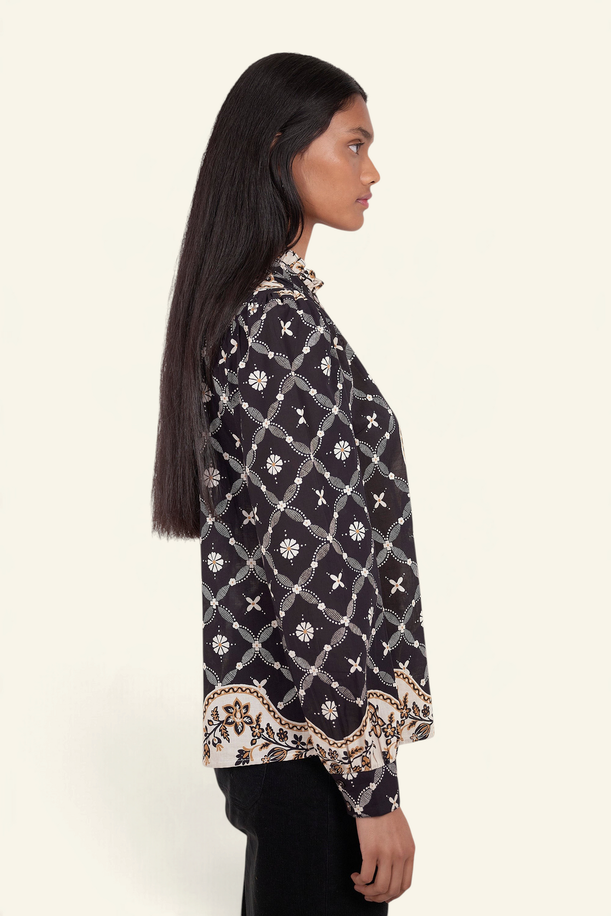 Side view of model standing. She is wearing Banjanan Christina shirt in black with a Victorian-inspired geometric bandana print, frilled collar, and puff sleeves.