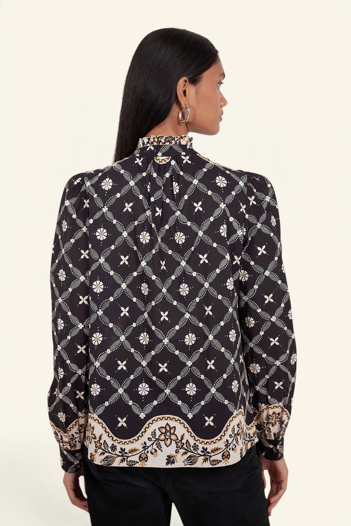 Back view of model standing. She is wearing Banjanan Christina shirt in black with a Victorian-inspired geometric bandana print, frilled collar, and puff sleeves.