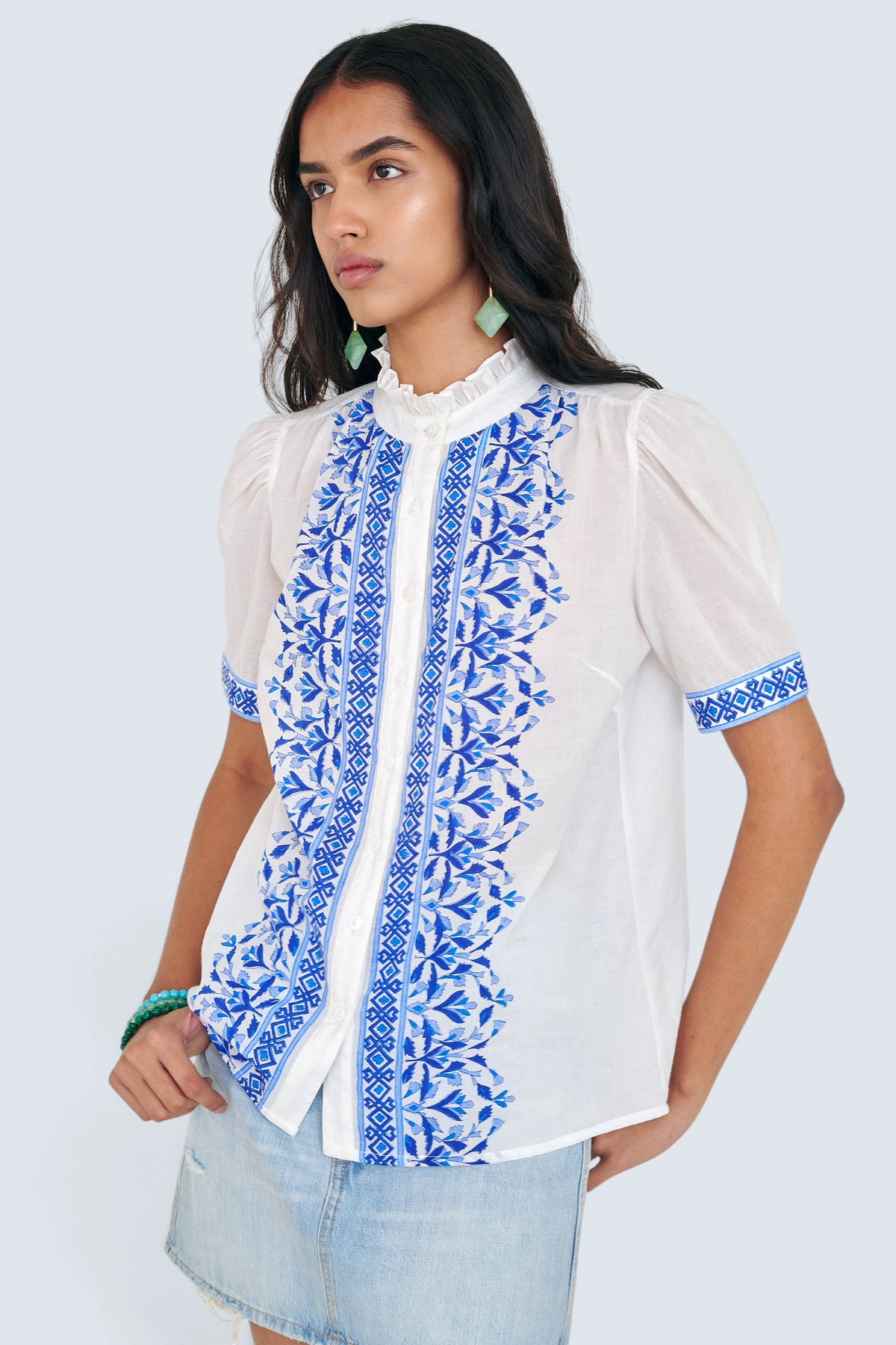 Front side view of woman standing. She is wearing the Christina shirt, featuring a hand block-printed geometric floral pattern, a frilled stand collar, soft gathers at the neckline, and slightly puffed sleeves. Paired with denim skirt.