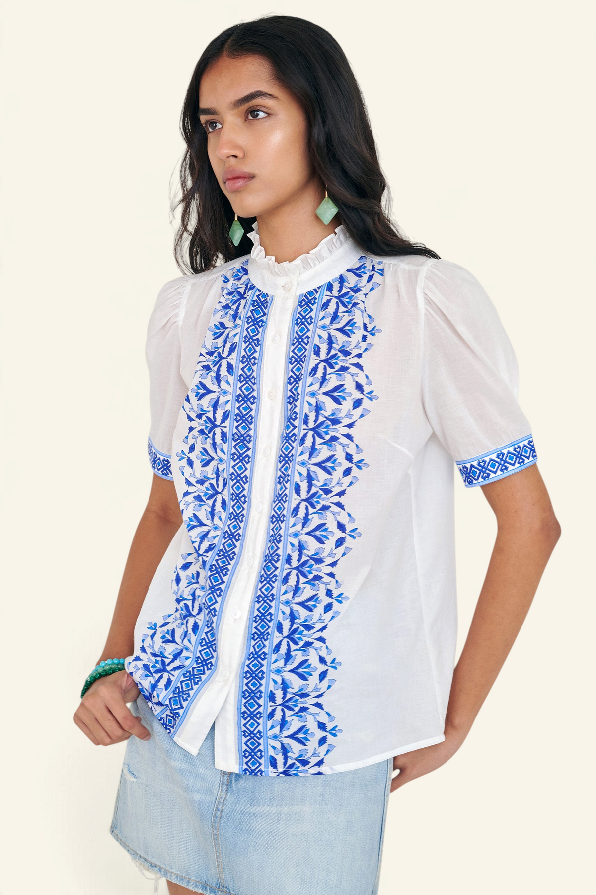 Front side view of woman standing. She is wearing the Christina shirt, featuring a hand block-printed geometric floral pattern, a frilled stand collar, soft gathers at the neckline, and slightly puffed sleeves. Paired with denim skirt.