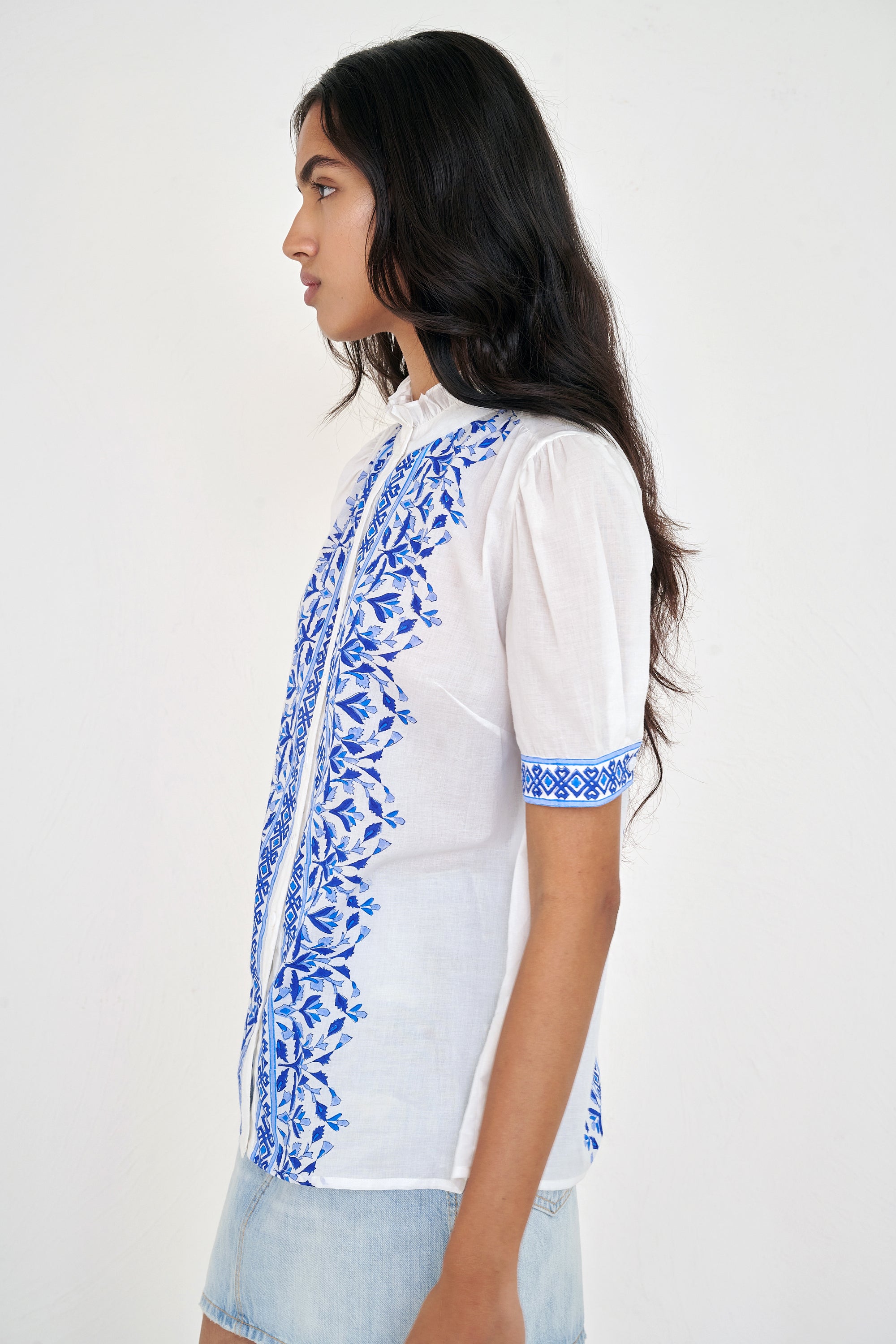 Side view of woman standing. She is wearing the Christina shirt, featuring a hand block-printed geometric floral pattern, a frilled stand collar, soft gathers at the neckline, and slightly puffed sleeves. Paired with denim skirt.