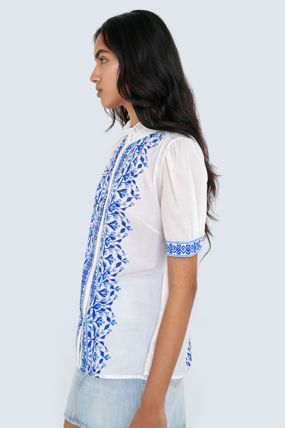Side view of woman standing. She is wearing the Christina shirt, featuring a hand block-printed geometric floral pattern, a frilled stand collar, soft gathers at the neckline, and slightly puffed sleeves. Paired with denim skirt.