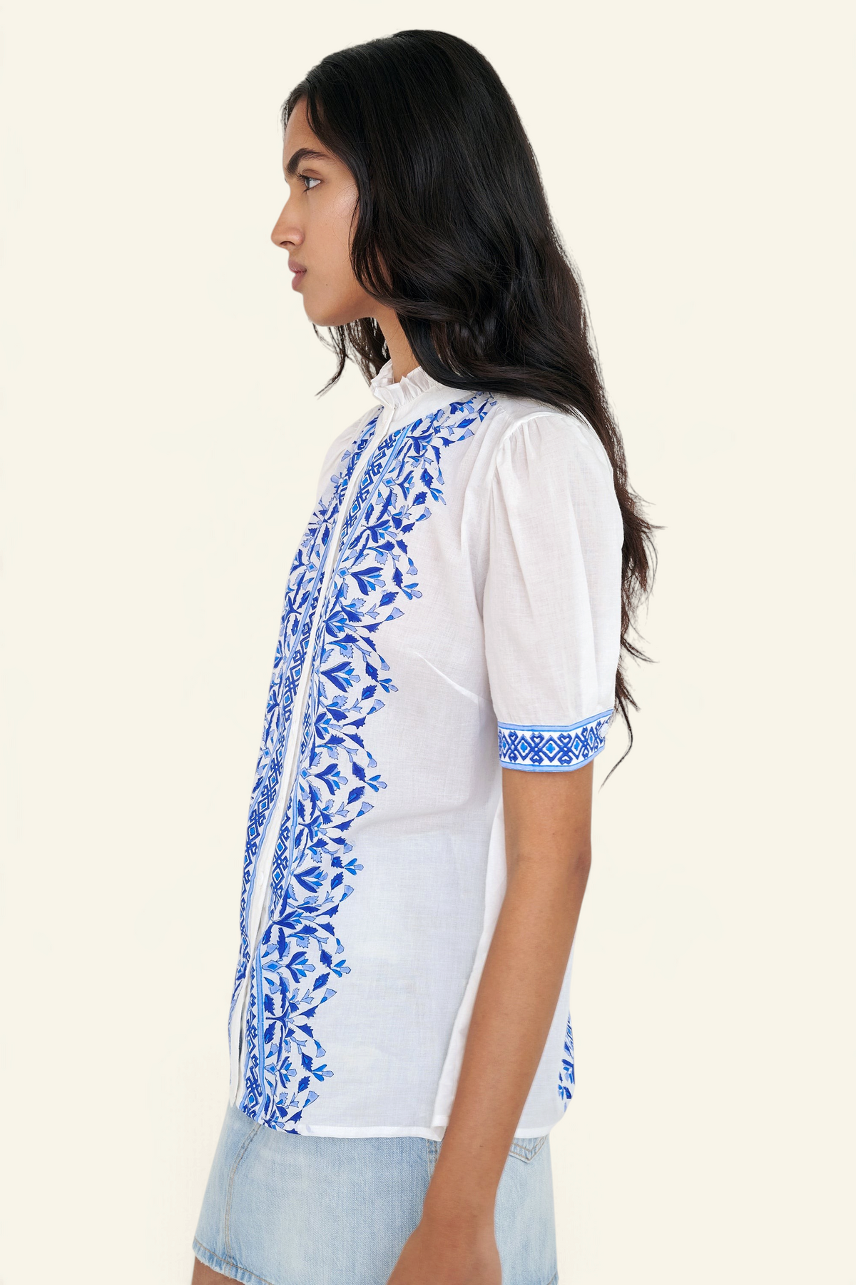 Side view of woman standing. She is wearing the Christina shirt, featuring a hand block-printed geometric floral pattern, a frilled stand collar, soft gathers at the neckline, and slightly puffed sleeves. Paired with denim skirt.