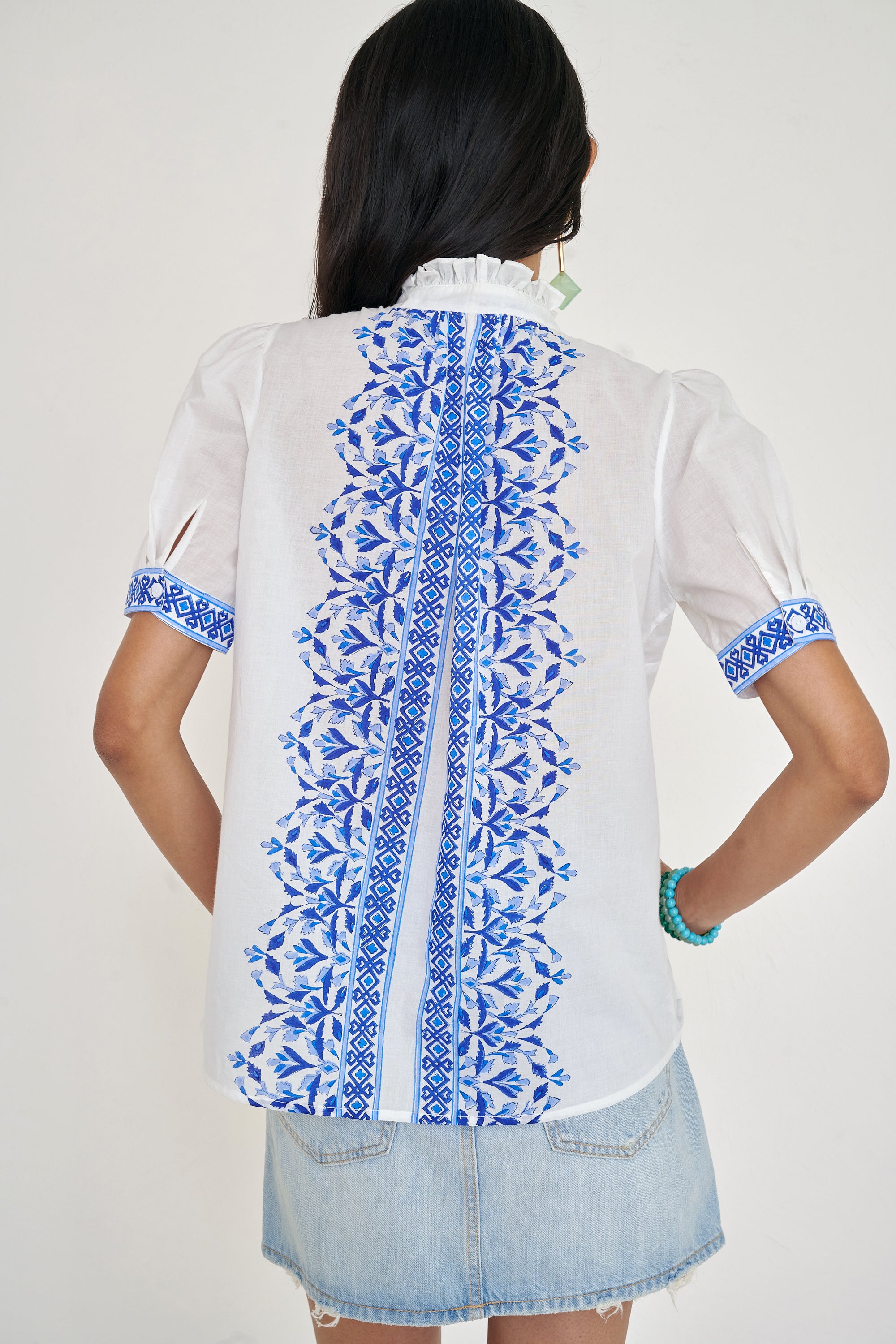 Back view of woman standing. She is wearing the Christina shirt, featuring a hand block-printed geometric floral pattern, a frilled stand collar, soft gathers at the neckline, and slightly puffed sleeves. Paired with denim skirt.