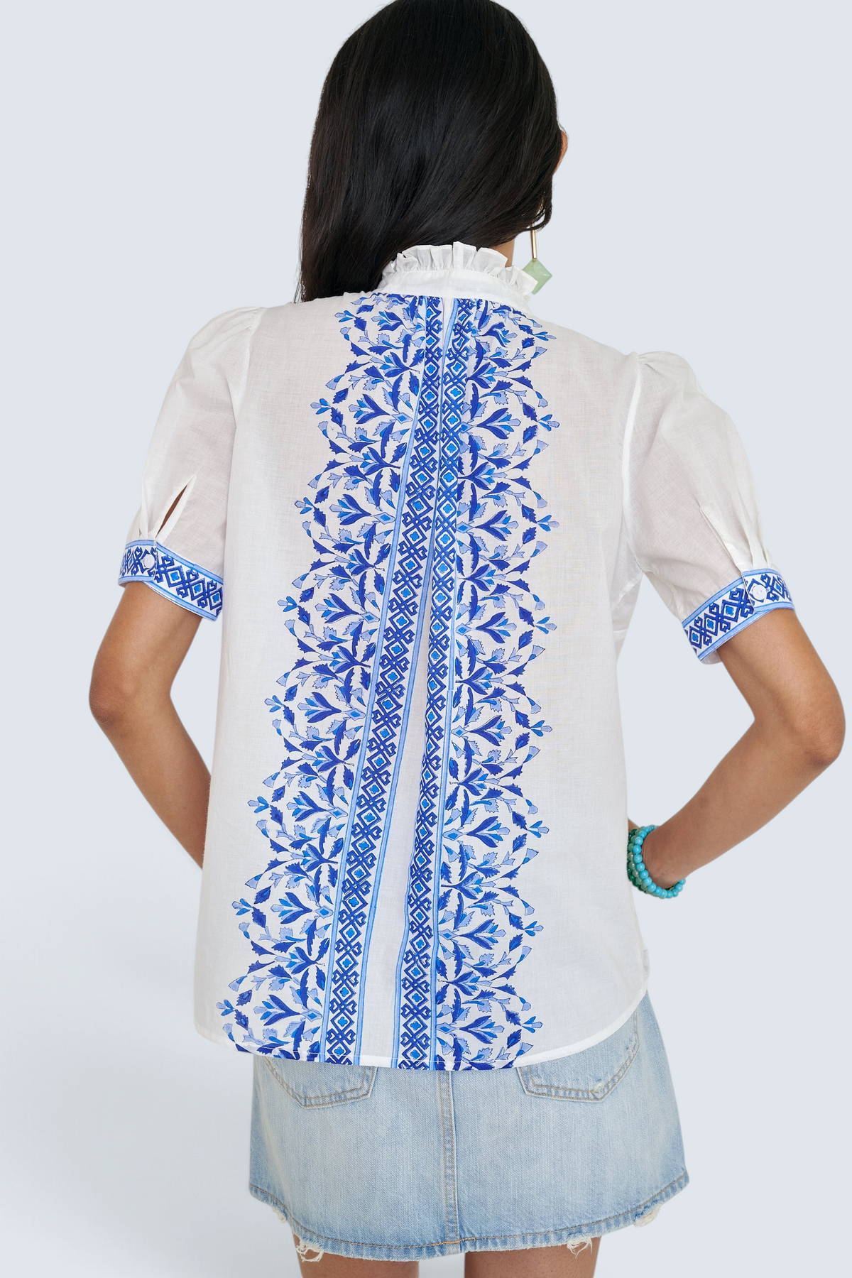 Back view of woman standing. She is wearing the Christina shirt, featuring a hand block-printed geometric floral pattern, a frilled stand collar, soft gathers at the neckline, and slightly puffed sleeves. Paired with denim skirt.