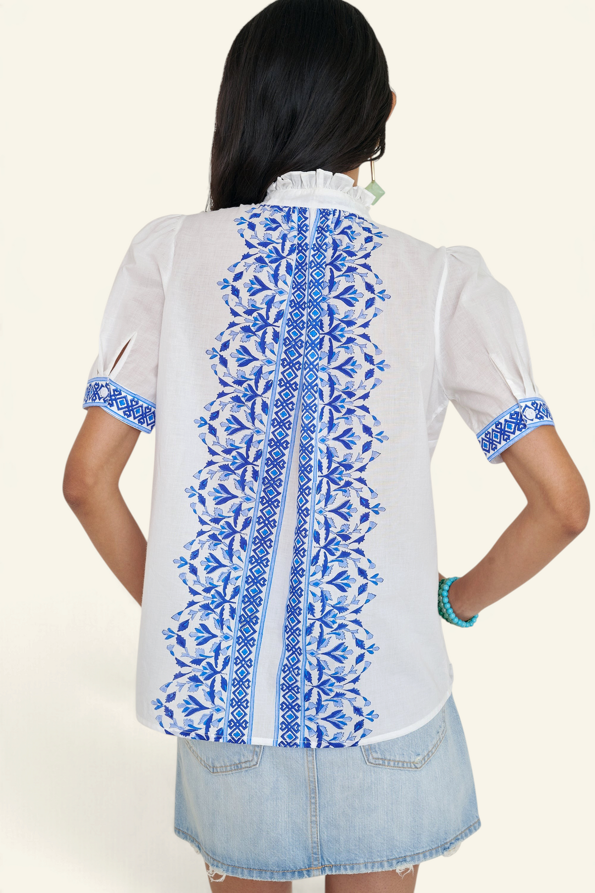 Back view of woman standing. She is wearing the Christina shirt, featuring a hand block-printed geometric floral pattern, a frilled stand collar, soft gathers at the neckline, and slightly puffed sleeves. Paired with denim skirt.