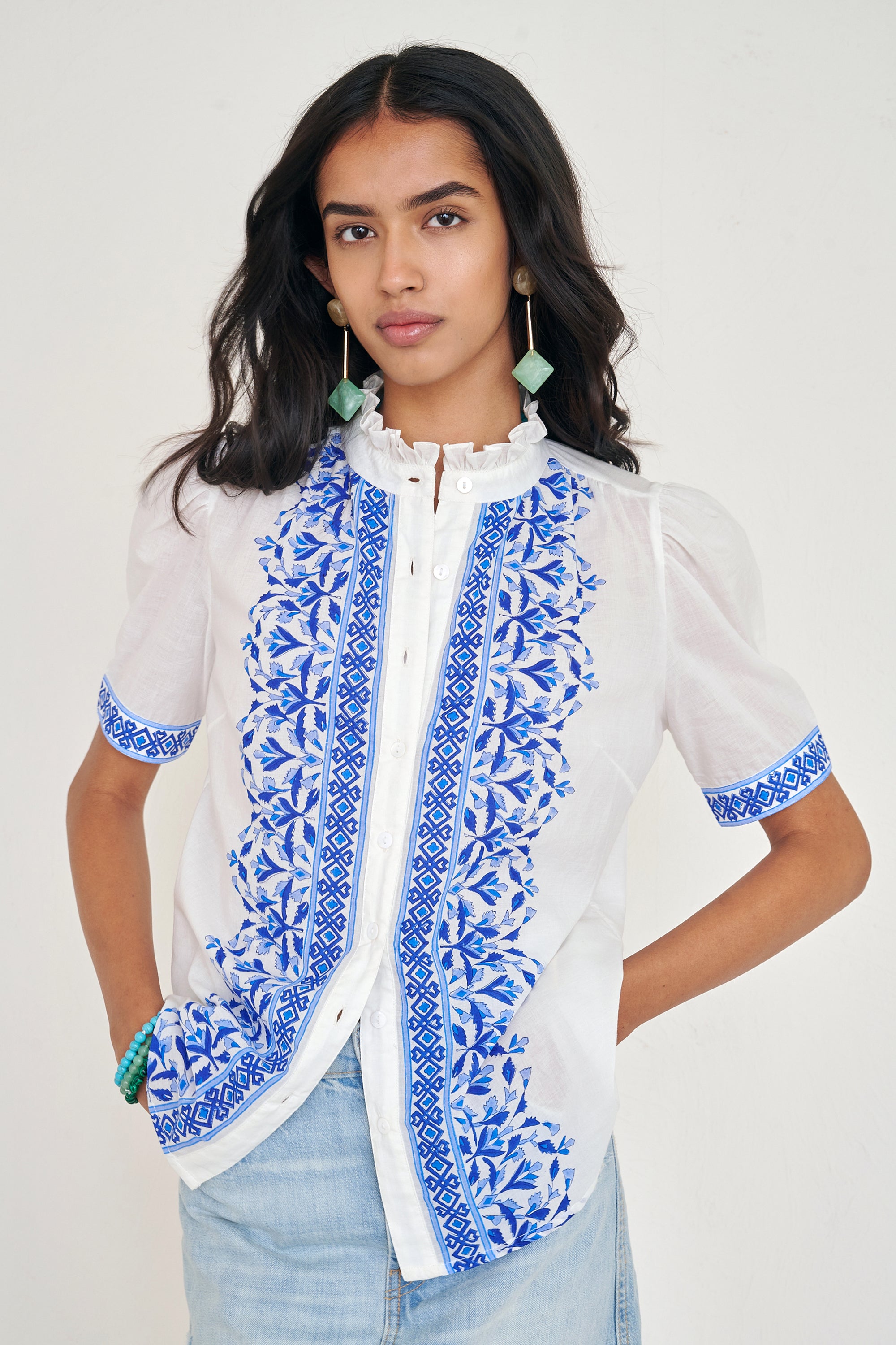 Front view of woman standing. She is wearing the Christina shirt, featuring a hand block-printed geometric floral pattern, a frilled stand collar, soft gathers at the neckline, and slightly puffed sleeves. Paired with denim skirt.