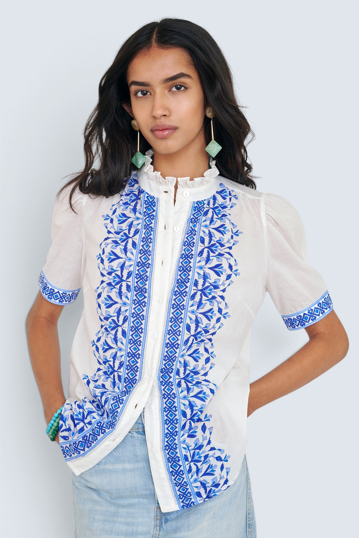 Front view of woman standing. She is wearing the Christina shirt, featuring a hand block-printed geometric floral pattern, a frilled stand collar, soft gathers at the neckline, and slightly puffed sleeves. Paired with denim skirt.