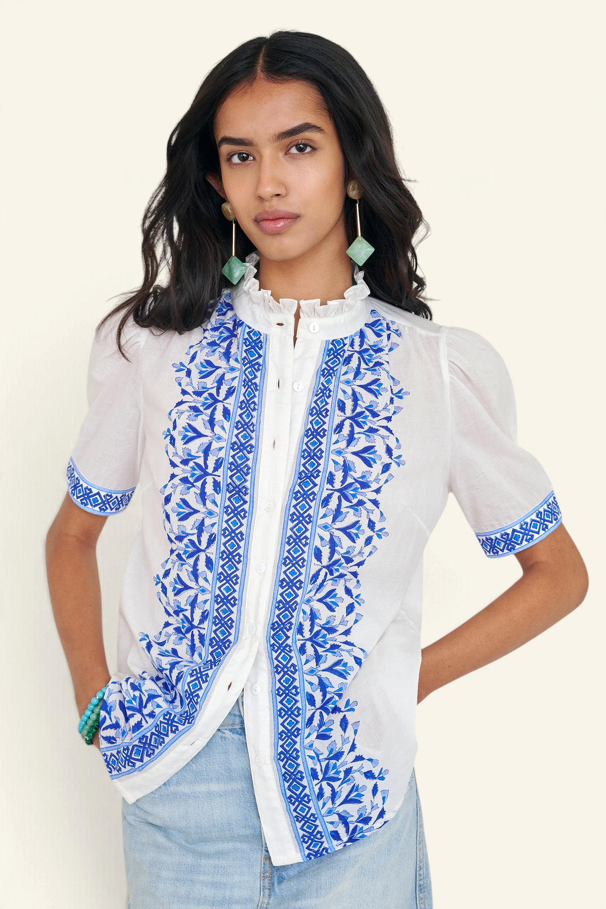 Front view of woman standing. She is wearing the Christina shirt, featuring a hand block-printed geometric floral pattern, a frilled stand collar, soft gathers at the neckline, and slightly puffed sleeves. Paired with denim skirt.