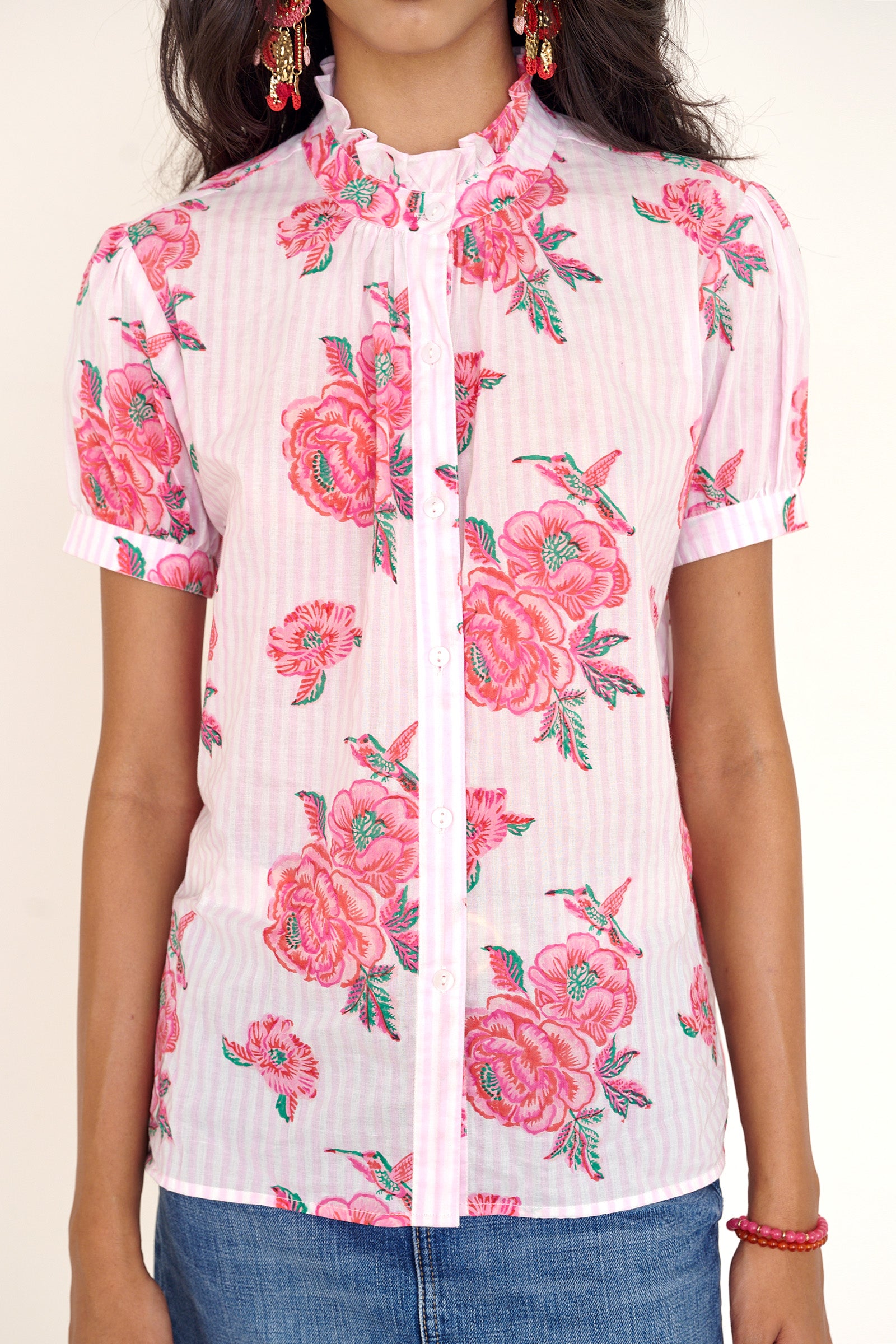 Front view of woman standing wearing the Christina shirt, featuring hand block-printed rose motifs, a ruffled neckline, subtly puffed short sleeves, and a button-front silhouette.