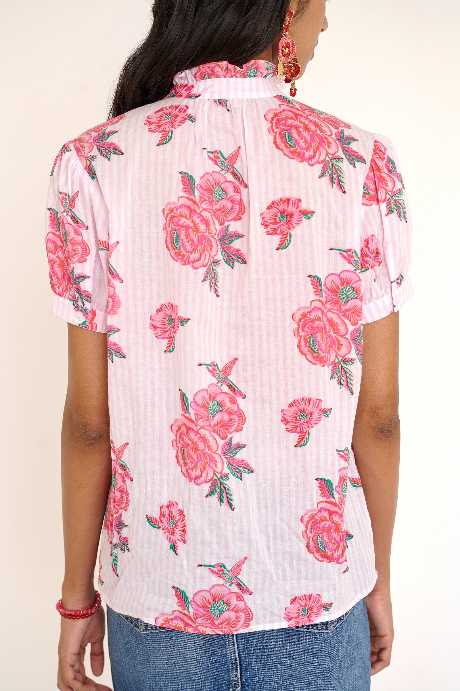 Back view of woman standing wearing the Christina shirt, featuring hand block-printed rose motifs, a ruffled neckline, subtly puffed short sleeves, and a button-front silhouette.