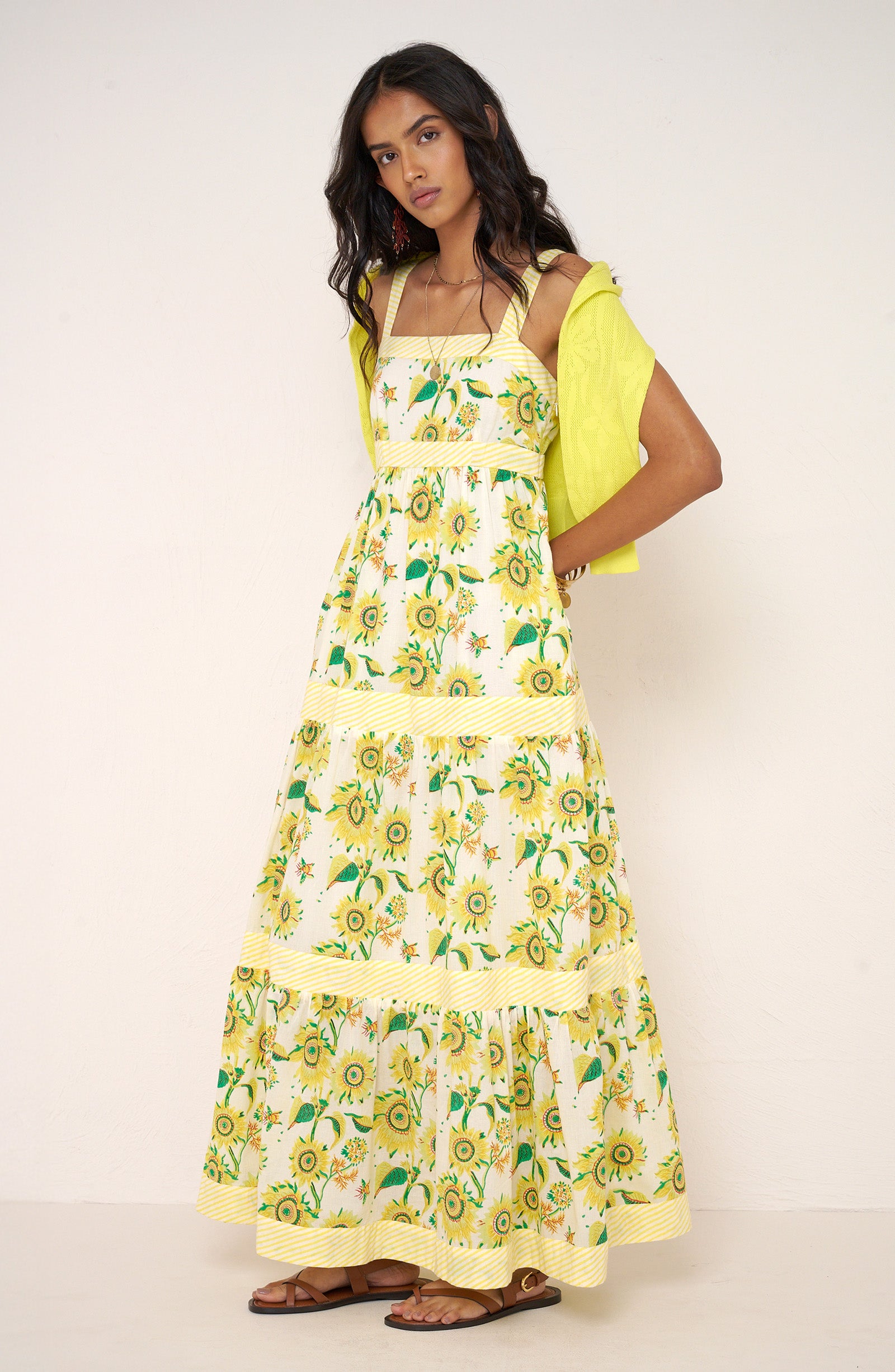 Side view of woman standing wearing the Daniella dress in the Sunflower print, featuring a flowing tiered silhouette, yellow stripe details, and hand-screen-printed sunflower motifs, styled with a citrus yellow cardigan draped over her shoulders.