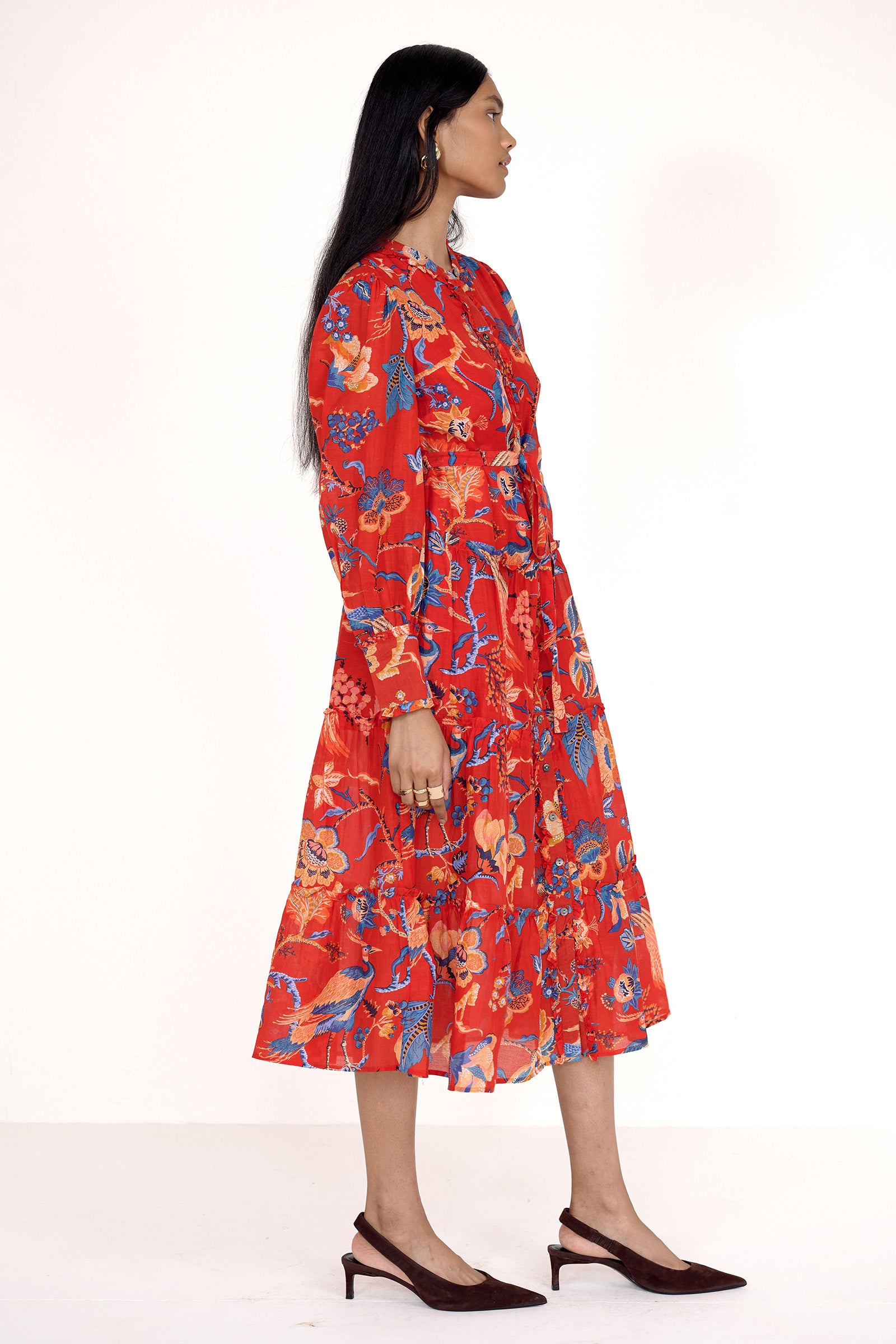 Side view of model standing. She is wearing the Emira dress in Chintz print. It features a flowing skirt, delicate ruffles, fabric-covered buttons, and a detachable belt.