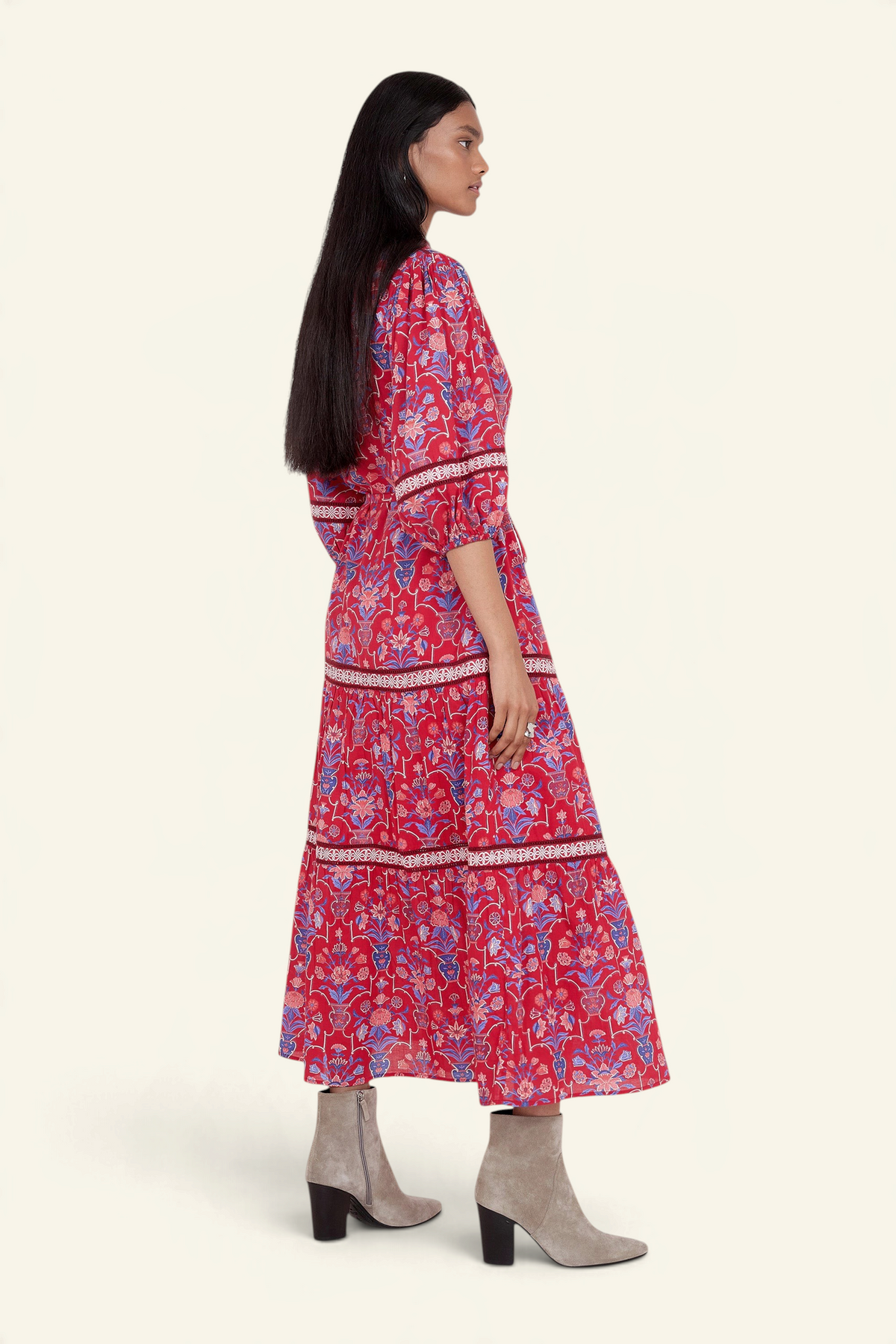 Side view of model standing. She is wearing the Liza dress in Floral Vase print. It features a tie neckline, soft ruffles, lace sleeve and skirt details, and a fabric belt.