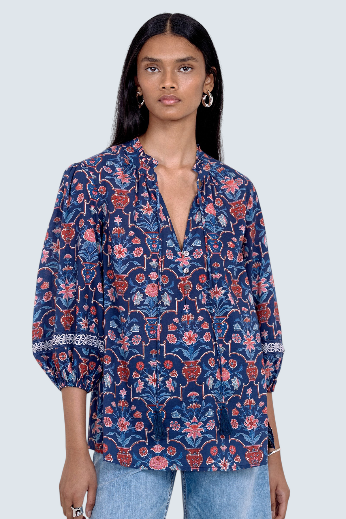 Front view of model standing. She is wearing the Lorna top in Floral Vase print, featuring a V-neck with a soft ruffle and tie, lace trim inset at the sleeves, a loose flowing body, and slightly voluminous sleeves, paired with jeans.