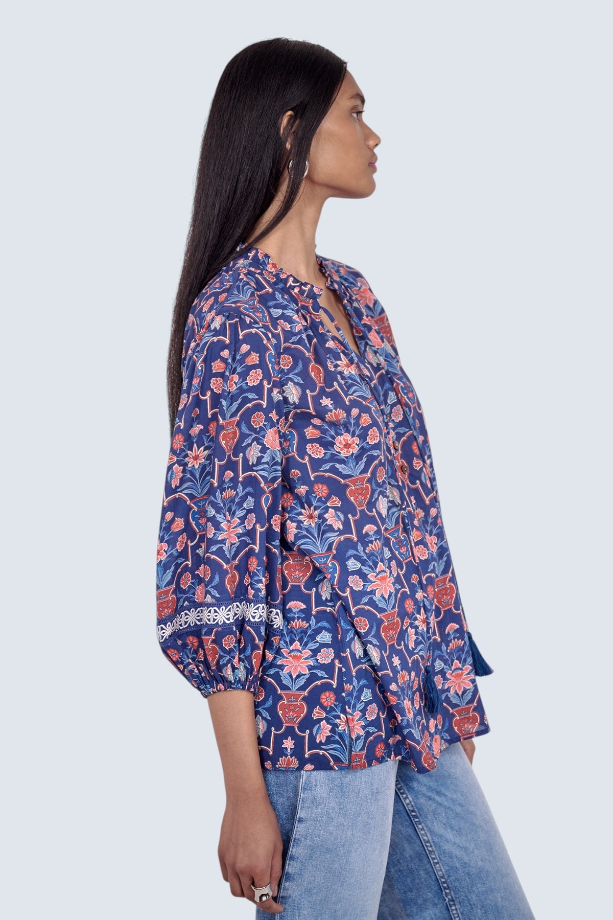 Side view of model standing wearing the Lorna top in Floral Vase print, featuring a V-neck with ruffle and tie, lace-trimmed sleeves, and a loose flowing silhouette, paired with jeans.