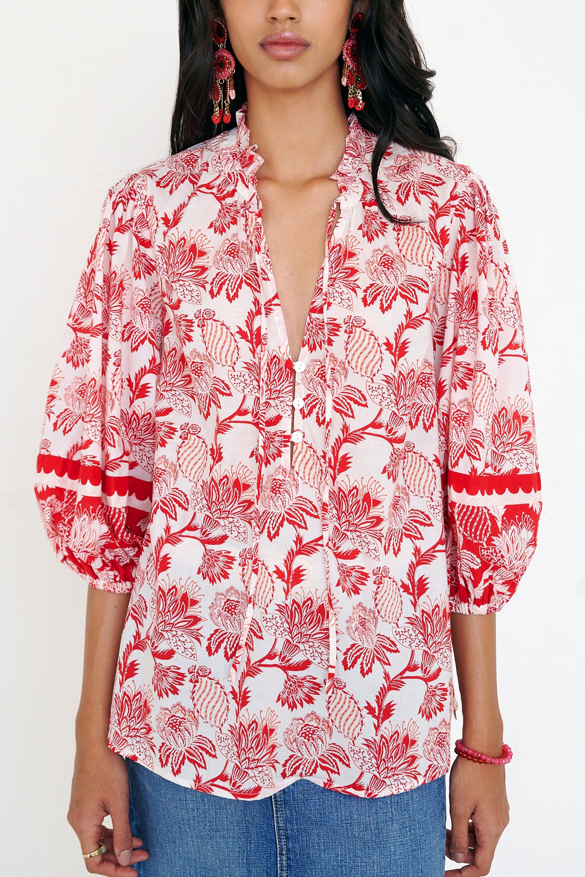 Front view of woman standing. She is wearing the Lorna top in the Lizzie print, featuring a V-neck with a soft ruffle, adjustable tie closure, voluminous sleeves, and winding floral vine motifs.