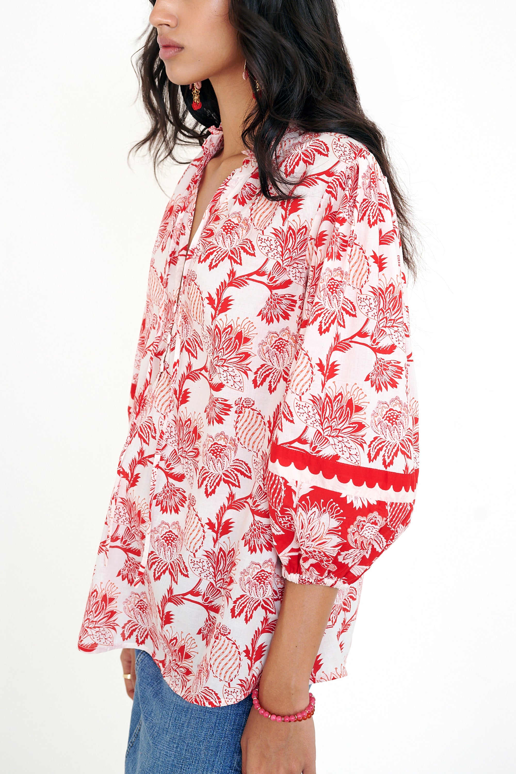 Side view of woman standing. She is wearing the Lorna top in the Lizzie print, featuring a V-neck with a soft ruffle, adjustable tie closure, voluminous sleeves, and winding floral vine motifs.