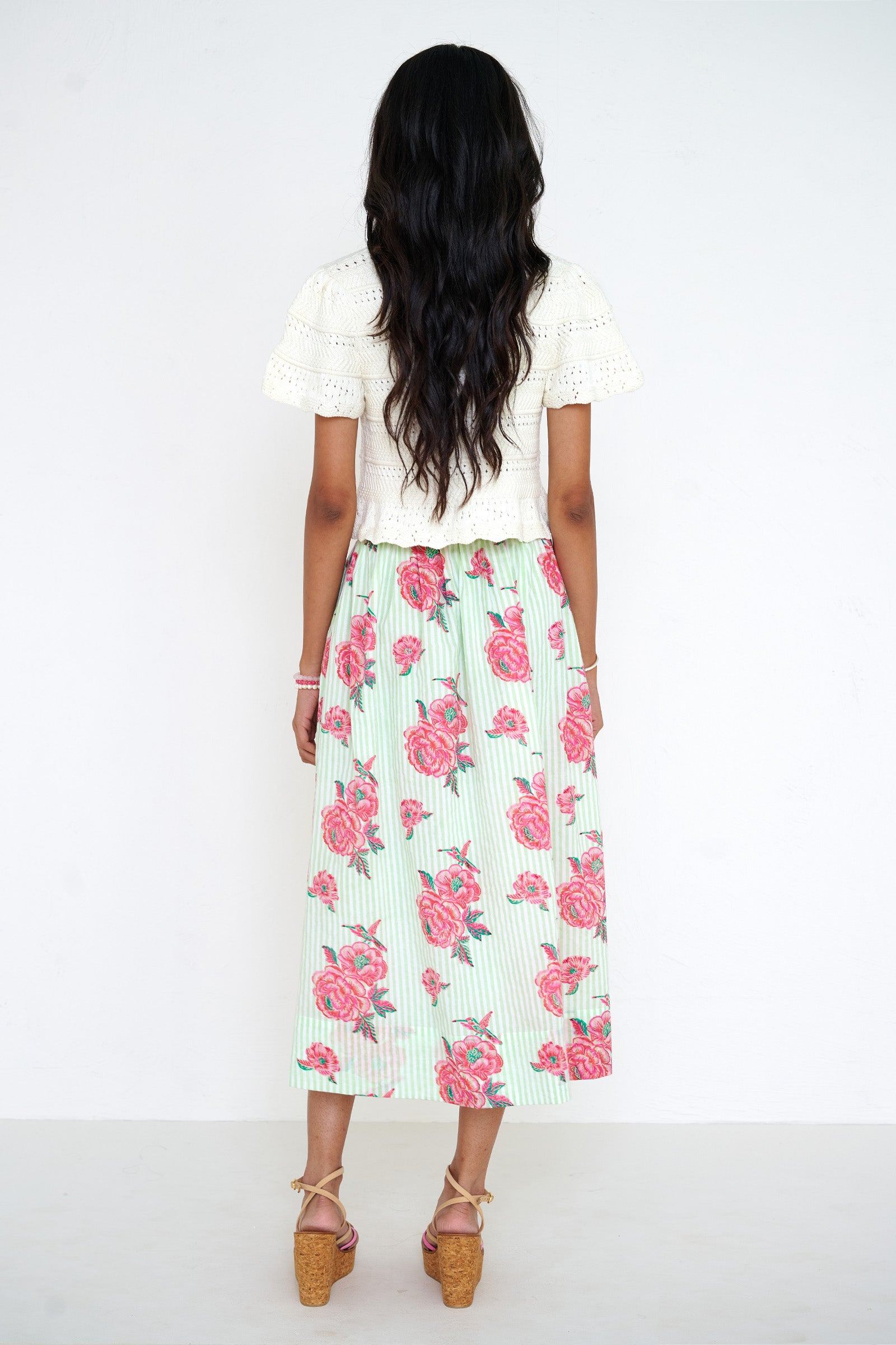 Back view of a woman standing wearing the Magdalena skirt in Eliza Rose print. It features all-around pleats, side pockets, and a side zipper closure with hand block-printed detailing. She paired it with our Laura crochet cardigan.
