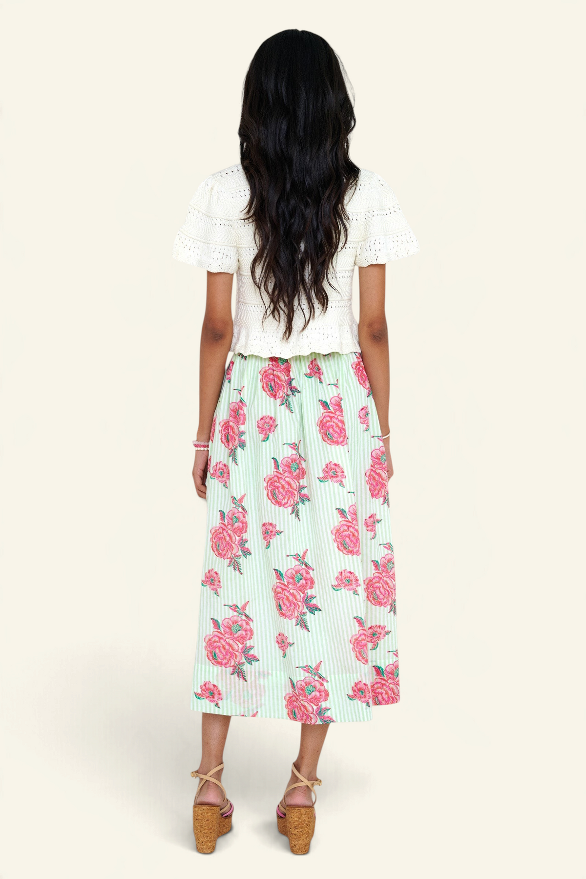 Back view of a woman standing wearing the Magdalena skirt in Eliza Rose print. It features all-around pleats, side pockets, and a side zipper closure with hand block-printed detailing. She paired it with our Laura crochet cardigan.