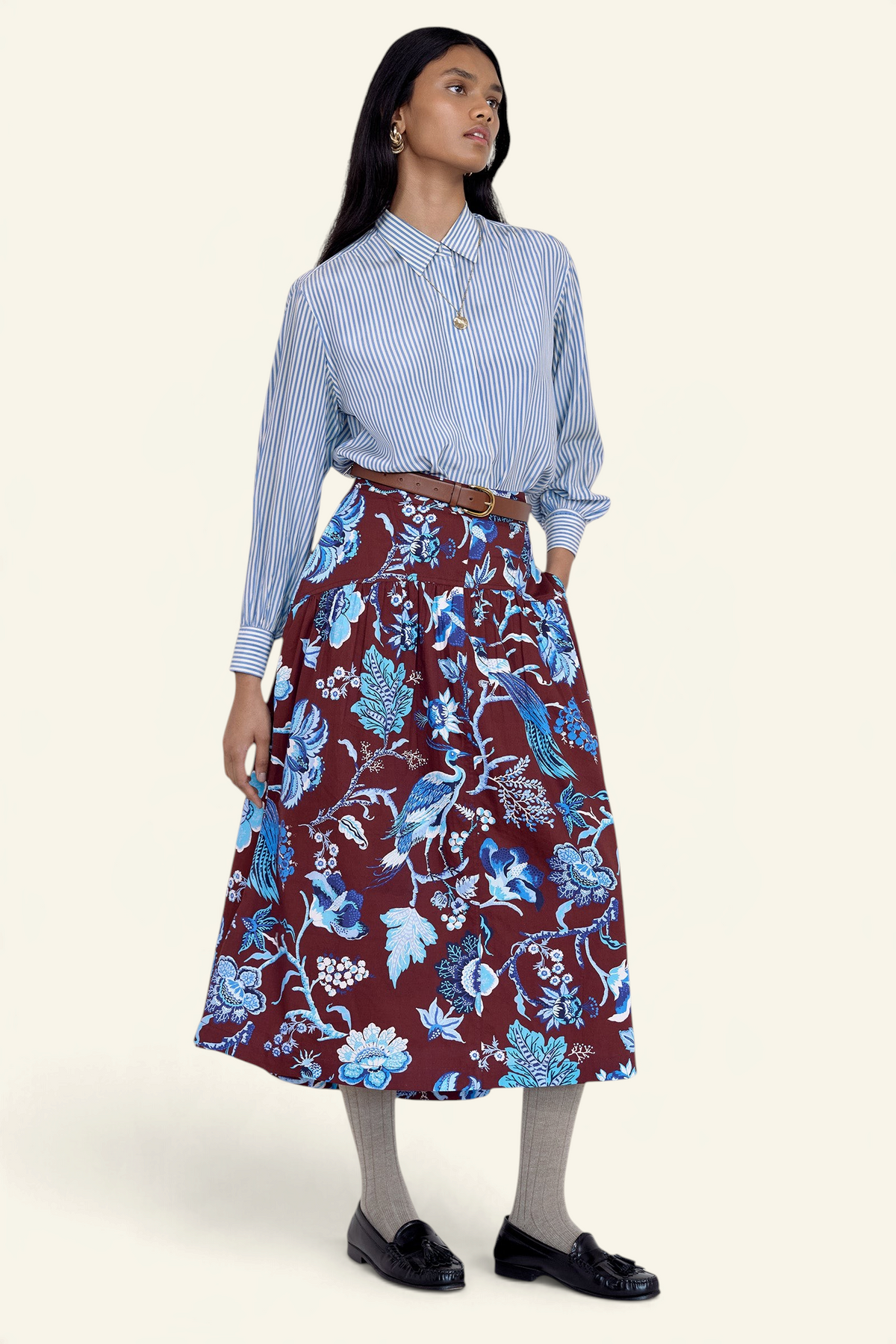 Front side view of model standing. She wearing the Melodie skirt in Chintz print, featuring a slim waist, gathered silhouette, and side-seam pockets.Its paired with a pinstripe shirt, belt, socks, shoe and accessories.