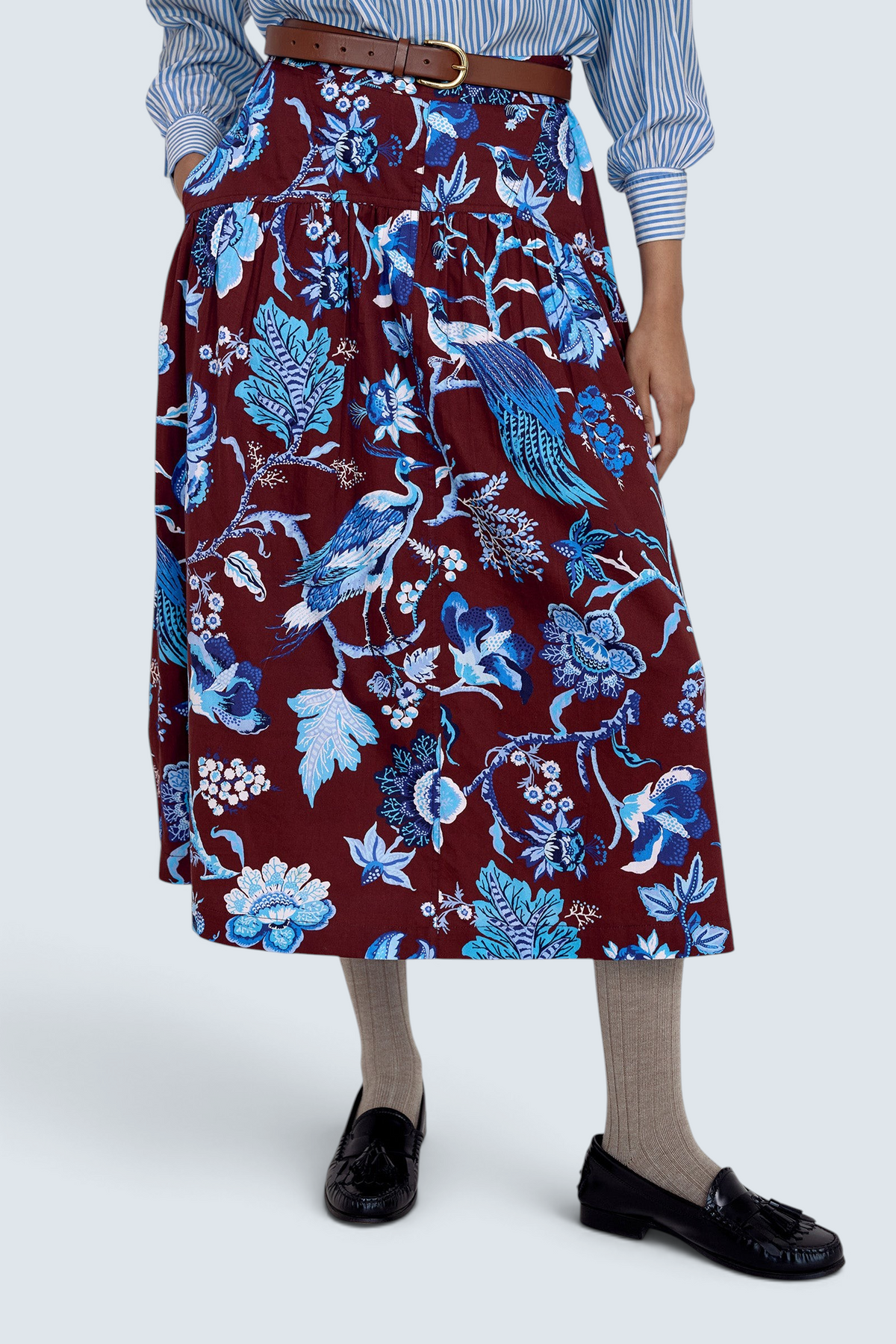 Front view close up of model standing. She wearing the Melodie skirt in Chintz print, featuring a slim waist, gathered silhouette, and side-seam pockets.Its paired with a pinstripe shirt, belt, socks, shoe and accessories.