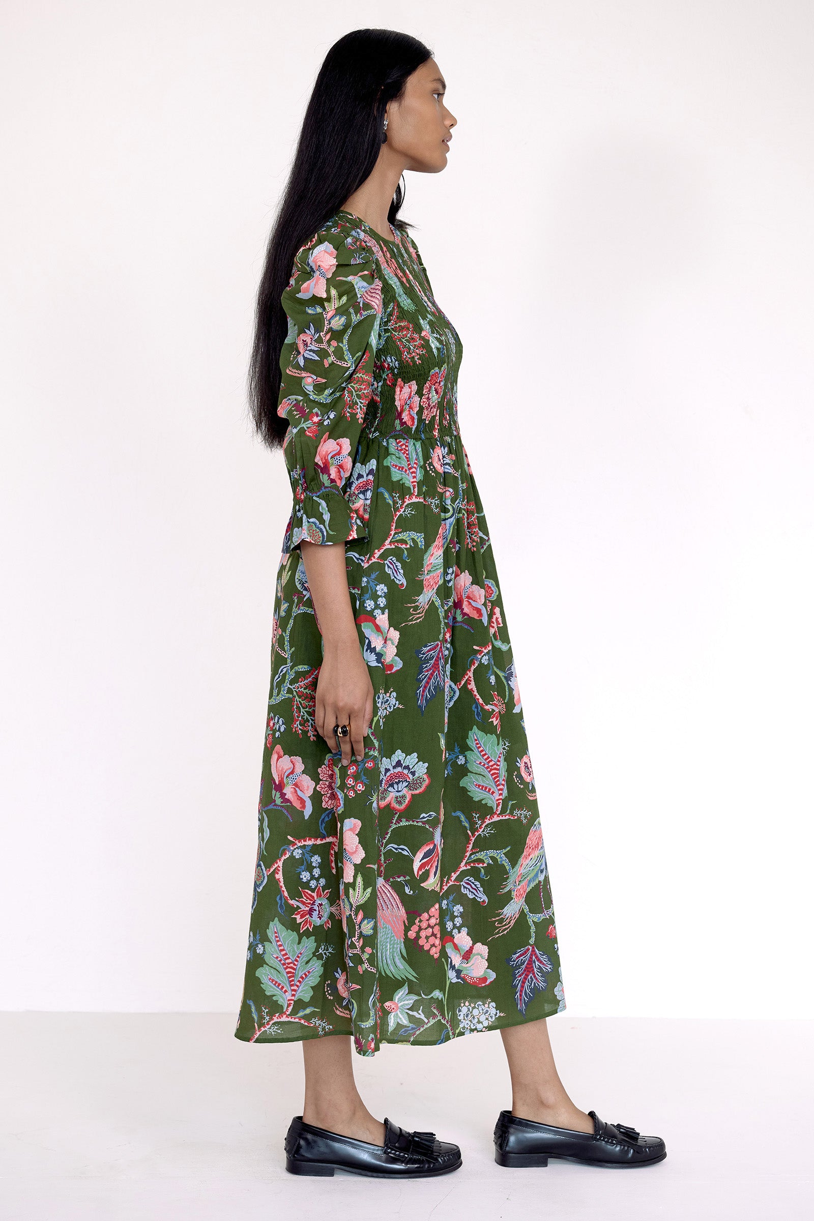 Side view of model standing wearing the Queenie dress in Chintz floral print. It features a smocked bodice, elbow-length puff sleeves, and a full skirt with pockets.