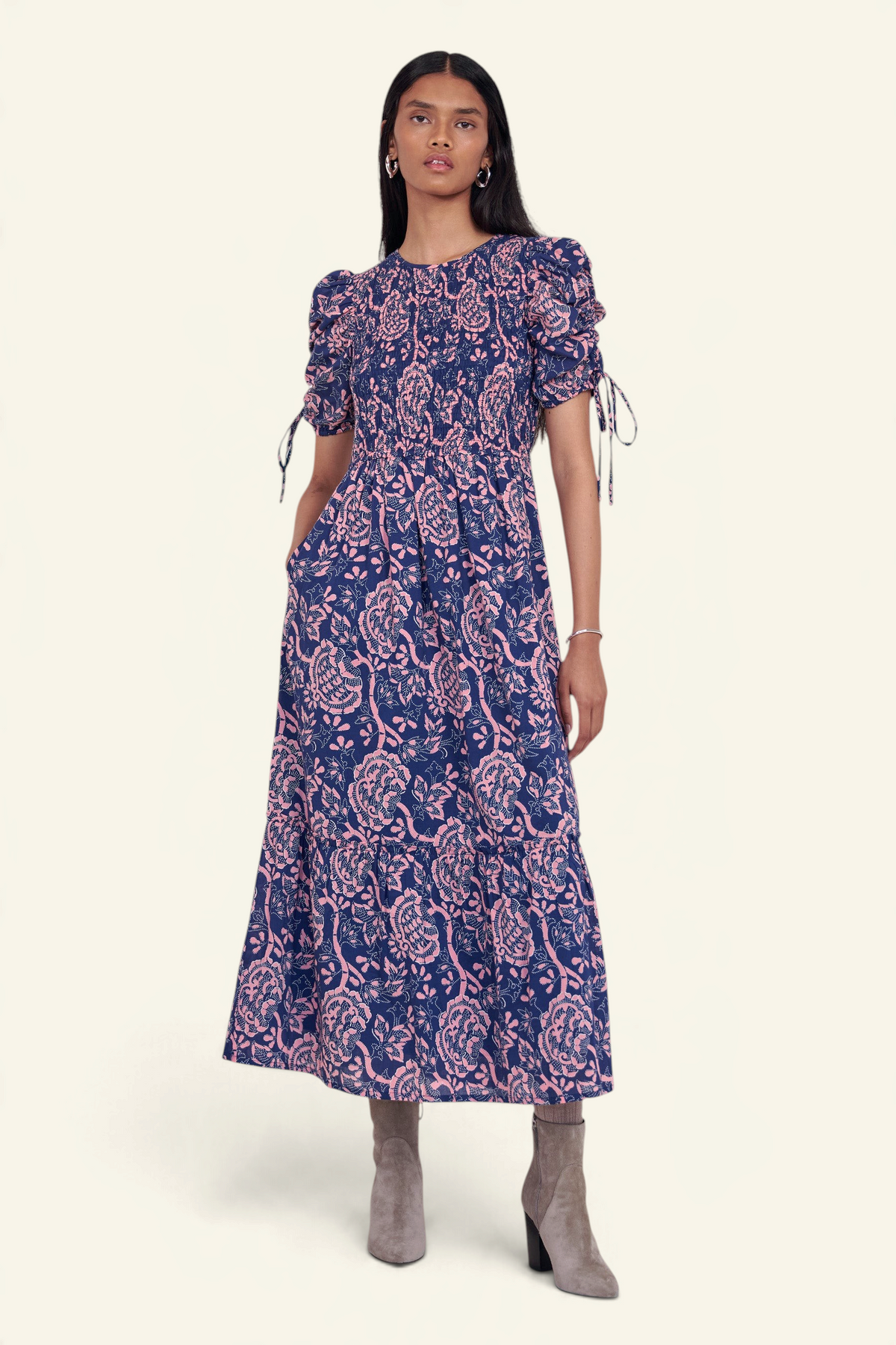 Front view of model standing. She is wearing the Queenie midi dress in Coromandel print, featuring a smocked bodice, adjustable drawstring sleeves, and a full lined skirt with pockets.