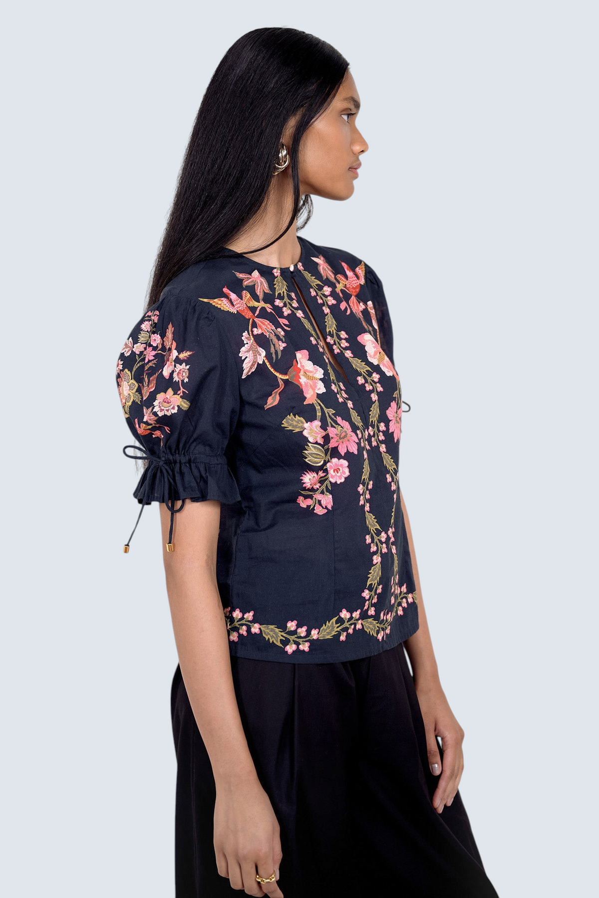 Side view of model standing. She is wearing the Ricki top in Chintz print, featuring a crewneck with keyhole detail, adjustable drawstring sleeves, and vintage-inspired print placement. Its paired with black pants.