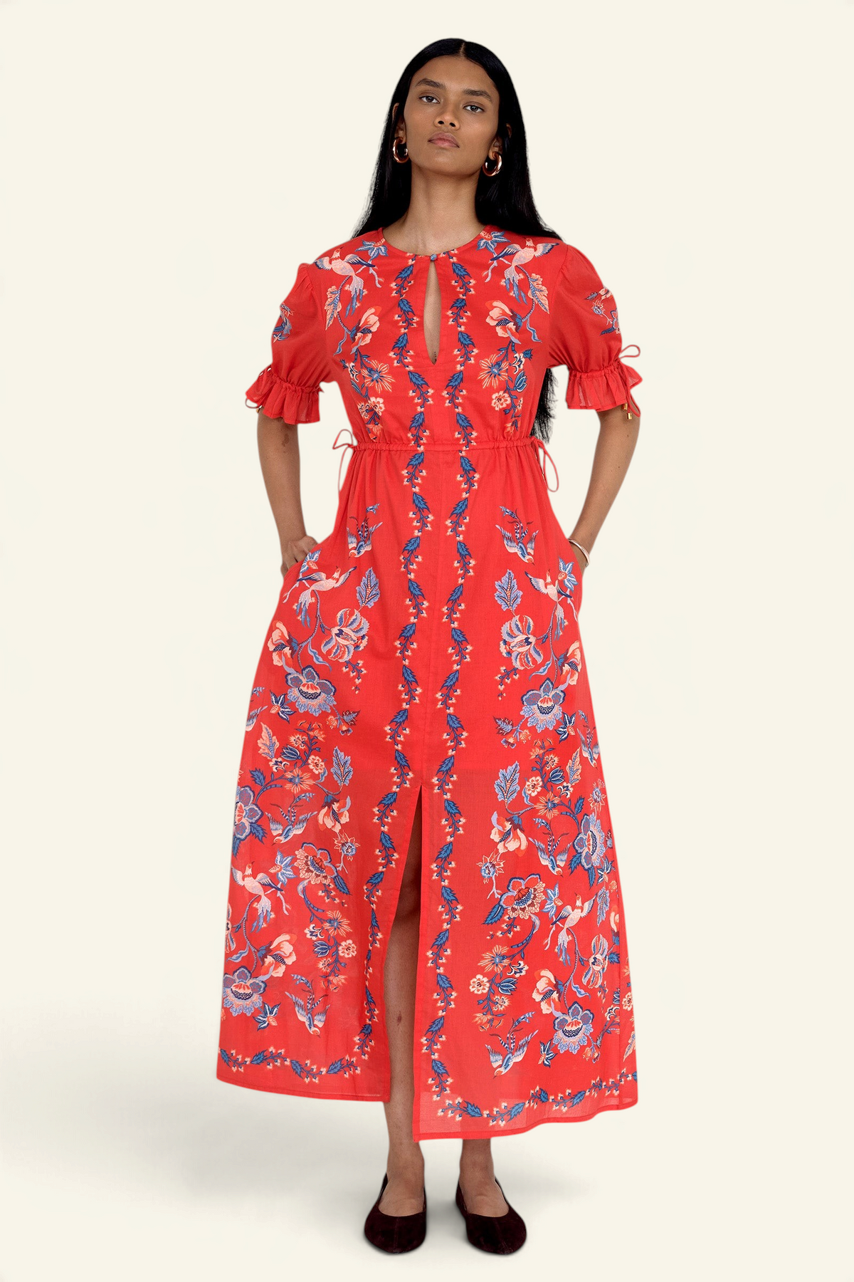 Front view of model standing. She is wearing the Ruby dress in Chintz print scarlet color with hands in pocket. It features a keyhole neckline, adjustable drawstring waist and sleeves, elbow-length sleeves, and an ankle-length hem.