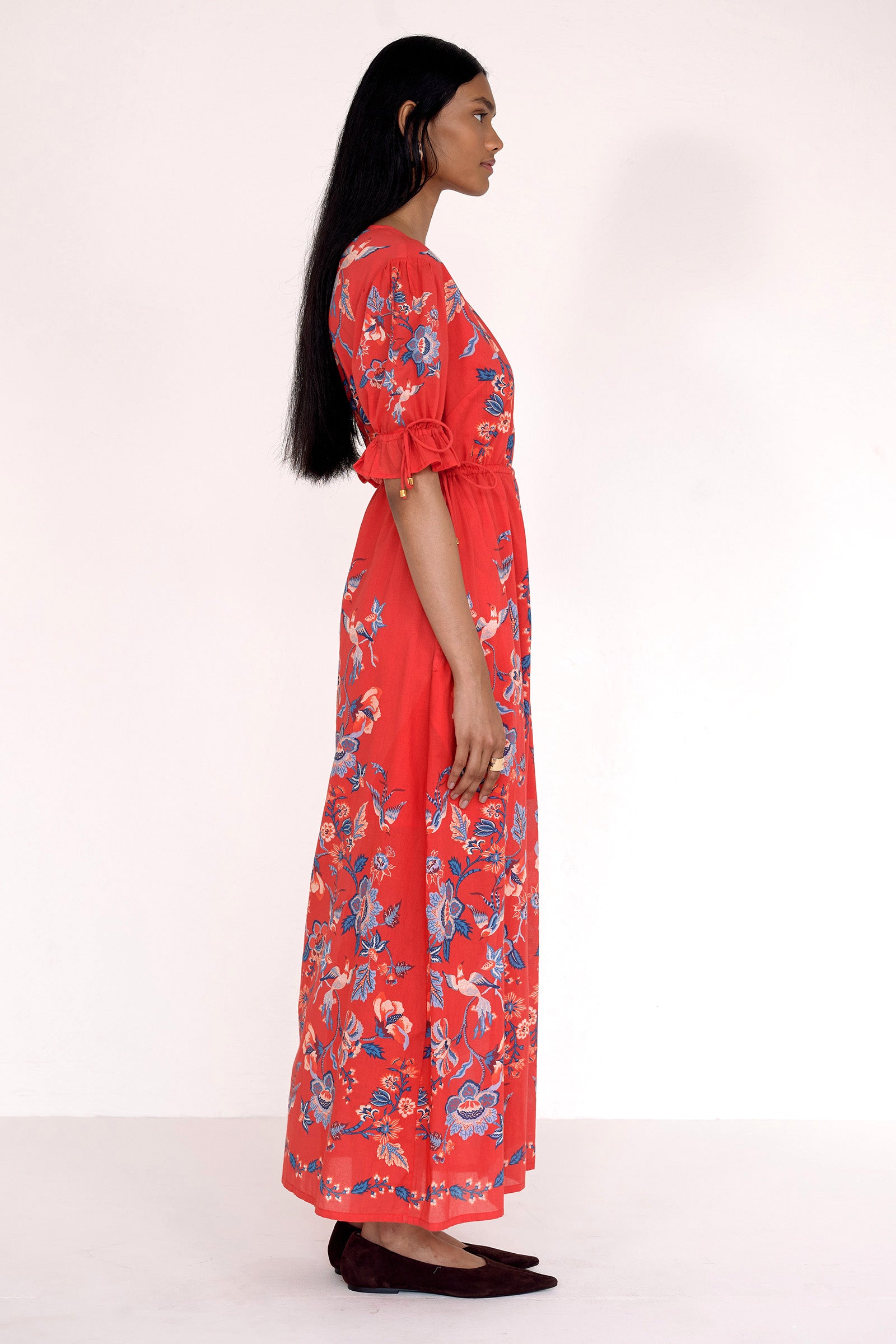 Side view of model standing. She is wearing the Ruby dress in Chintz print scarlet color. It features a keyhole neckline, adjustable drawstring waist and sleeves, elbow-length sleeves, and an ankle-length hem.