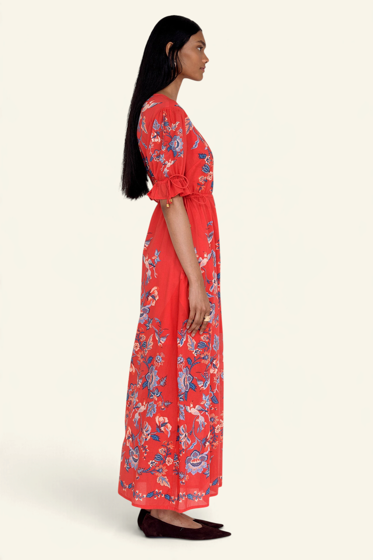 Side view of model standing. She is wearing the Ruby dress in Chintz print scarlet color. It features a keyhole neckline, adjustable drawstring waist and sleeves, elbow-length sleeves, and an ankle-length hem.