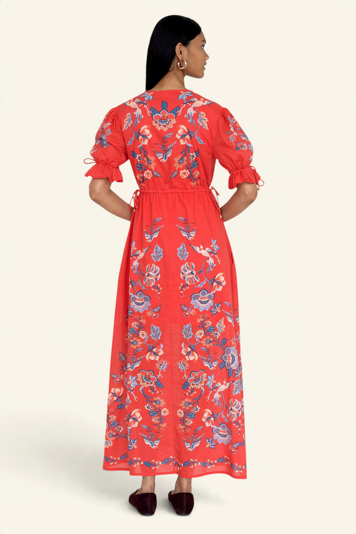 Back view of model standing. She is wearing the Ruby dress in Chintz print scarlet color. It features a keyhole neckline, adjustable drawstring waist and sleeves, elbow-length sleeves, and an ankle-length hem.