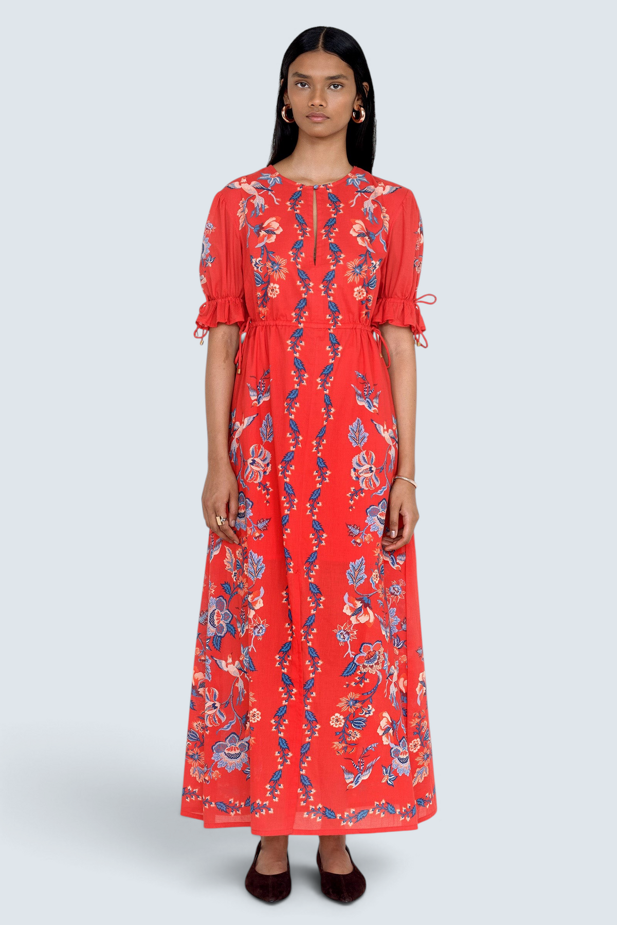 Front view of model standing. She is wearing the Ruby dress in Chintz print scarlet color. It features a keyhole neckline, adjustable drawstring waist and sleeves, elbow-length sleeves, and an ankle-length hem.
