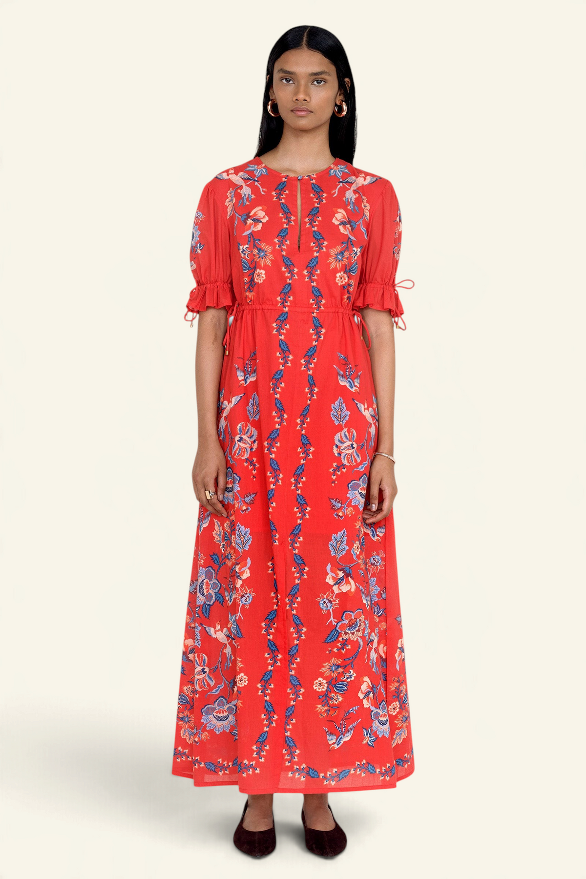 Front view of model standing. She is wearing the Ruby dress in Chintz print scarlet color. It features a keyhole neckline, adjustable drawstring waist and sleeves, elbow-length sleeves, and an ankle-length hem.