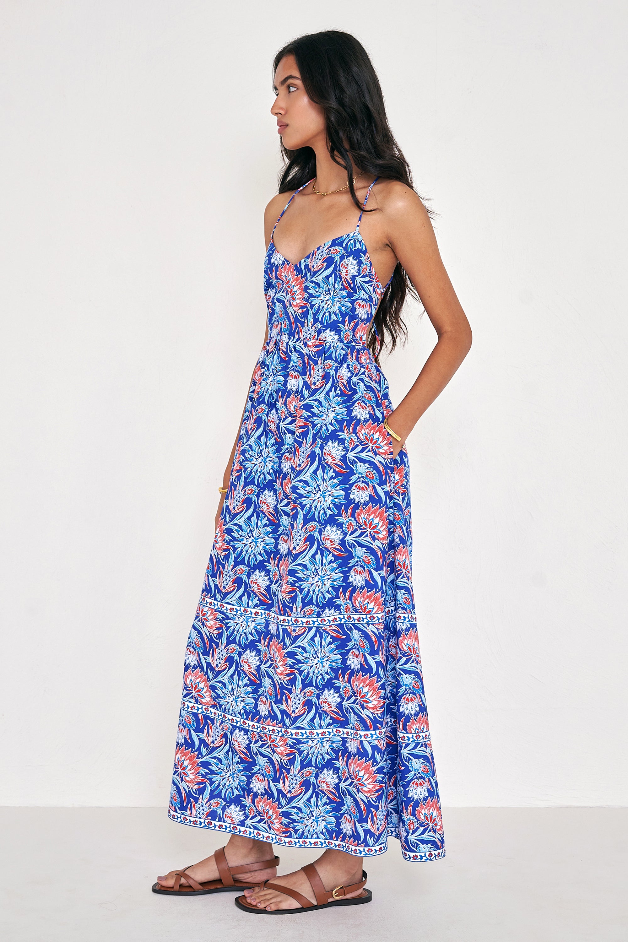 Side view of woman standing. She is wearing the Sofia dress in the Philippa print, featuring a strappy sundress silhouette, gathered ankle-length skirt, and red, white, and blue wildflower motifs inspired by Provence.