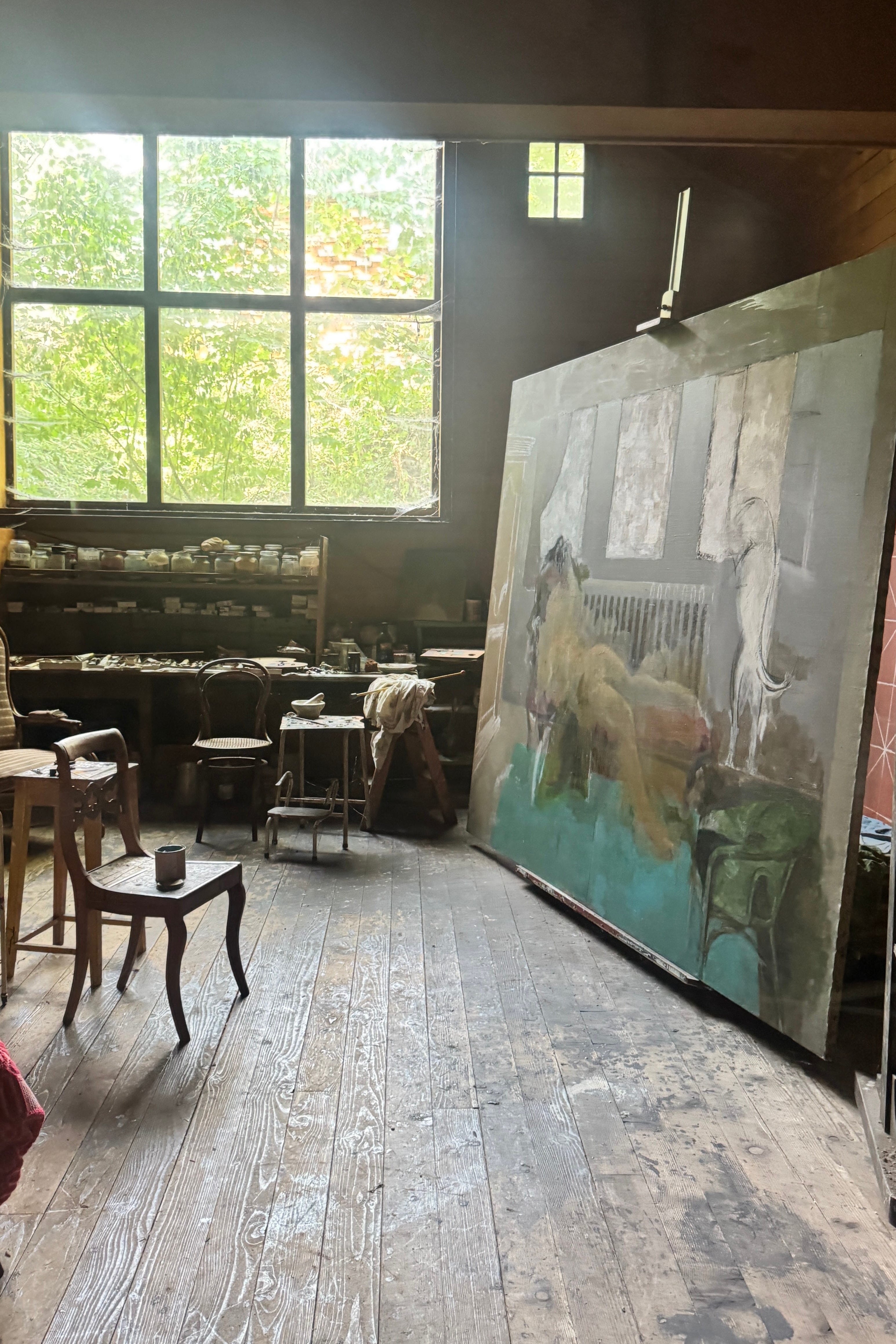 A large canvas painting of a horse is displayed in a rustic artist studio with wooden floors, work tables, and a large window showing green trees outside.