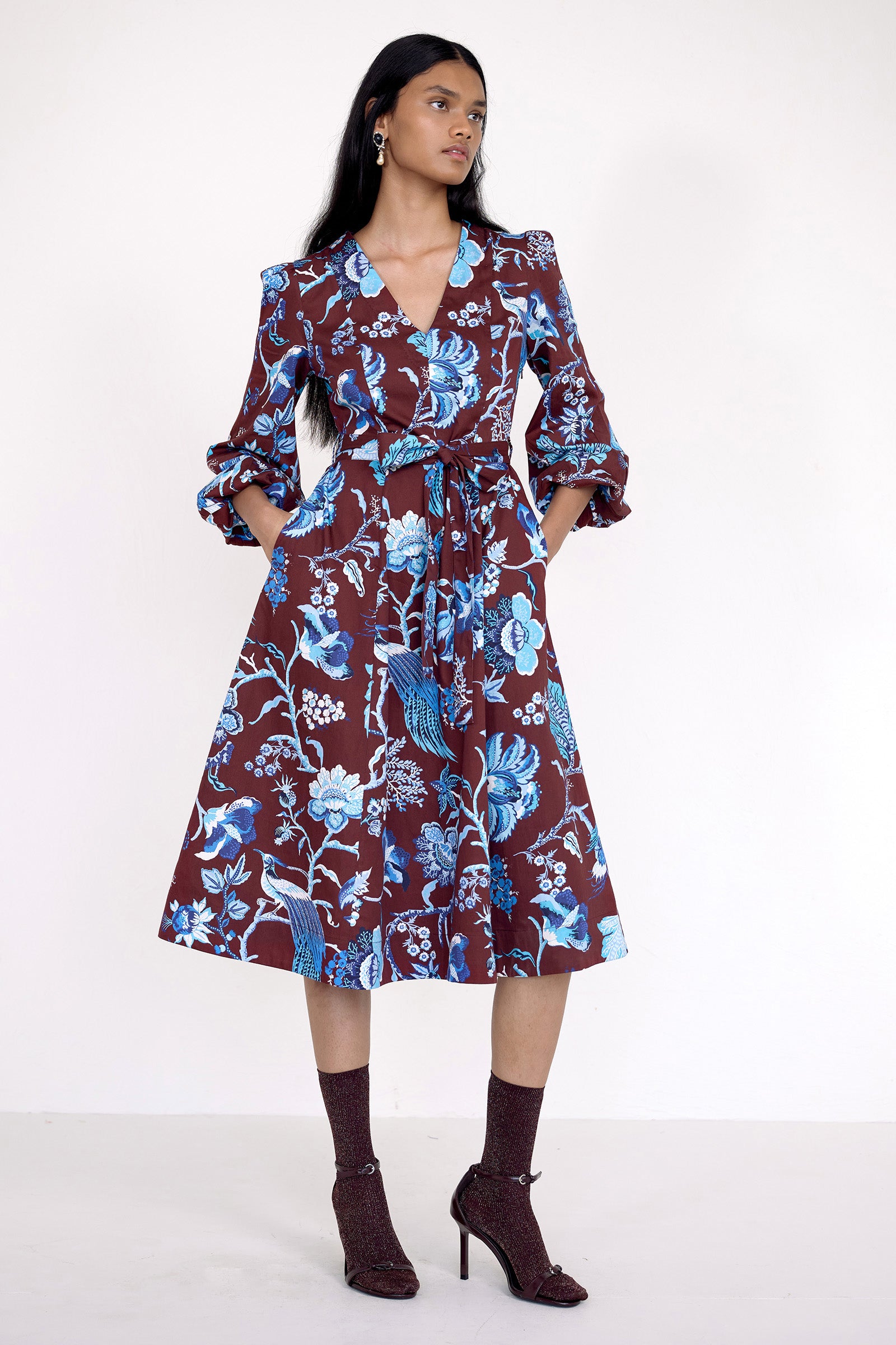 Front view of model standing. She is wearing the Zora dress in Chintz print, featuring a V-neck, lantern sleeves, princess seams, a gathered fit-and-flare skirt, fabric belt, and side-seam pockets.