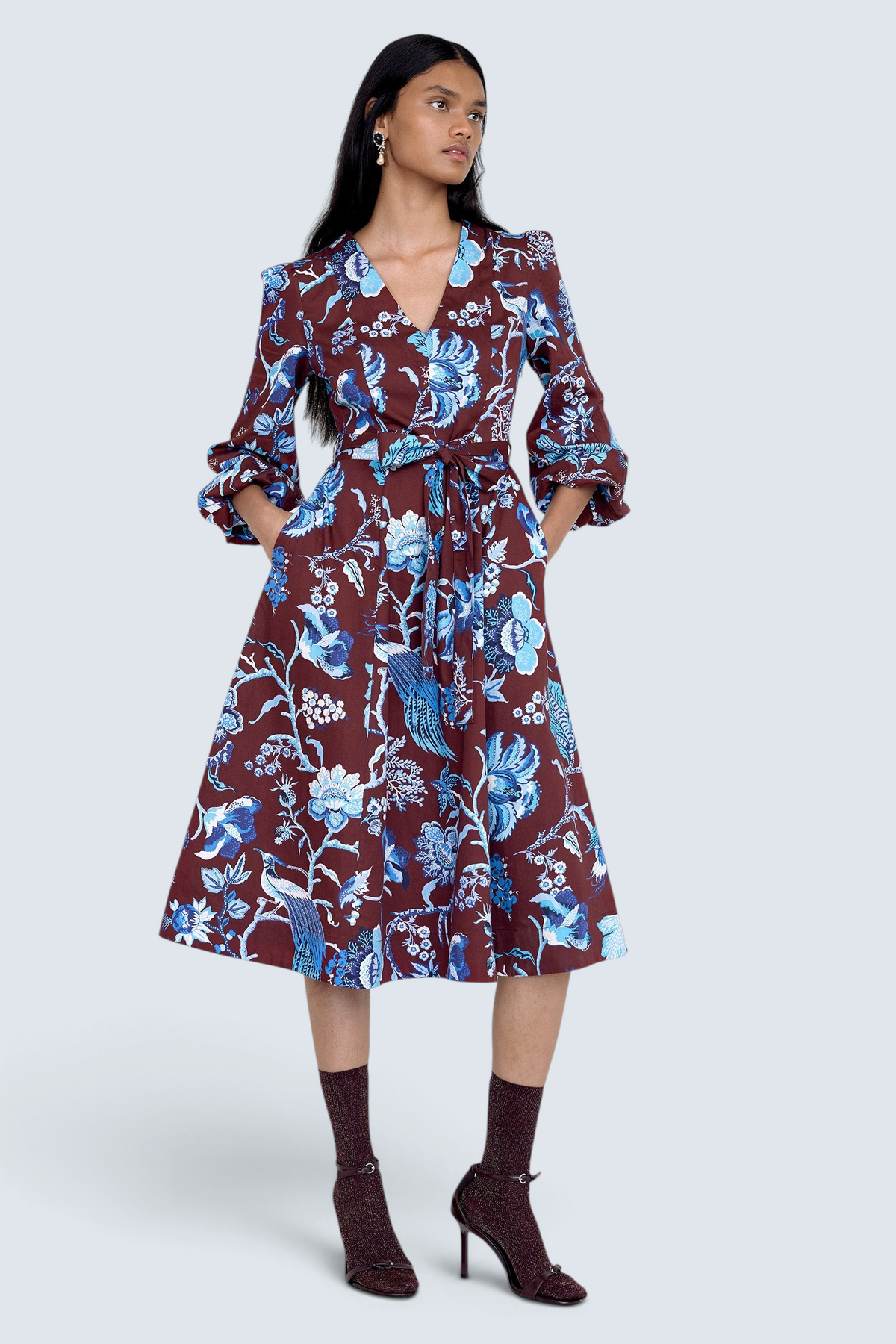 Front view of model standing. She is wearing the Zora dress in Chintz print, featuring a V-neck, lantern sleeves, princess seams, a gathered fit-and-flare skirt, fabric belt, and side-seam pockets.