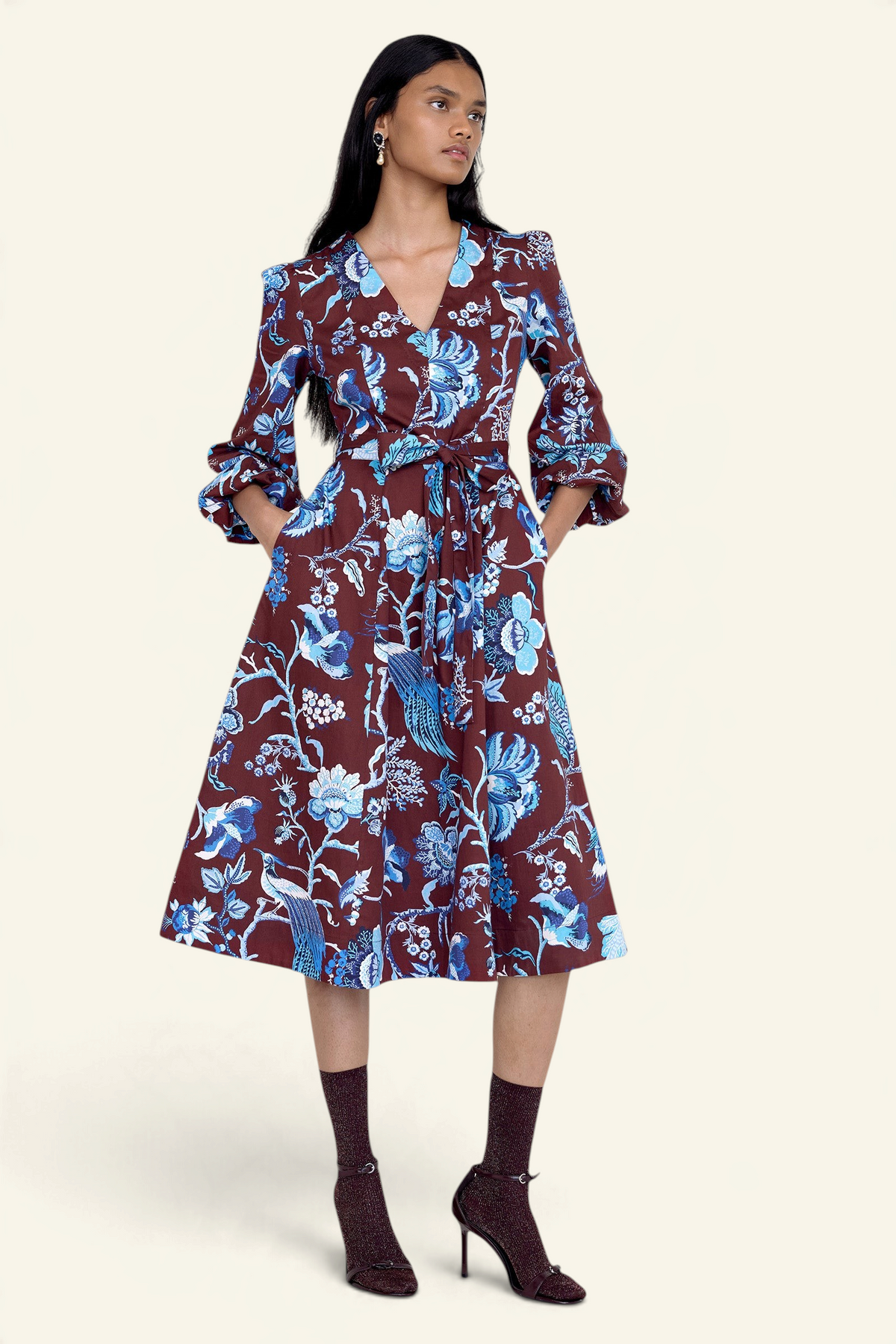 Front view of model standing. She is wearing the Zora dress in Chintz print, featuring a V-neck, lantern sleeves, princess seams, a gathered fit-and-flare skirt, fabric belt, and side-seam pockets.