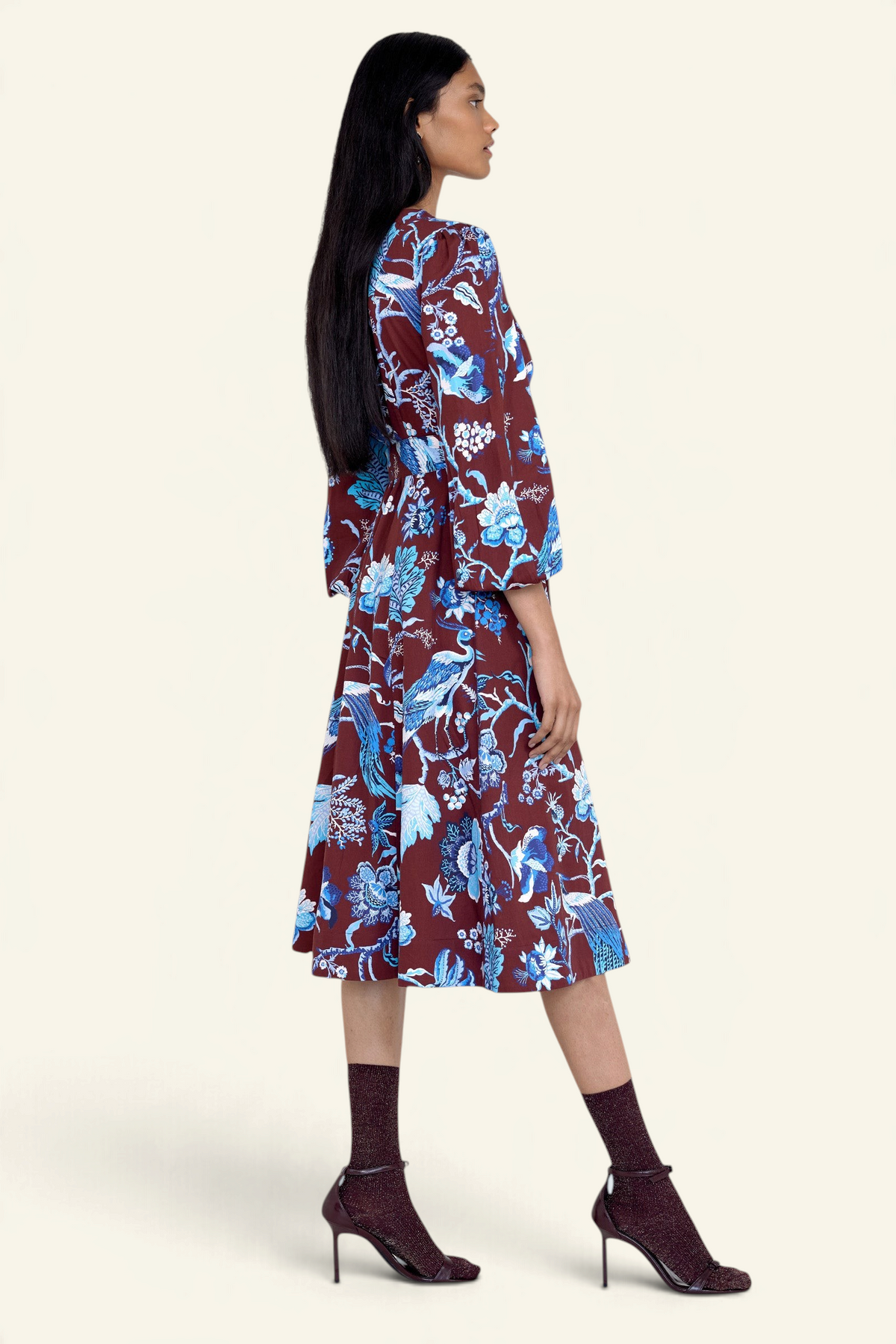 Side view of model standing. She is wearing the Zora dress in Chintz print, featuring a V-neck, lantern sleeves, princess seams, a gathered fit-and-flare skirt, fabric belt, and side-seam pockets.