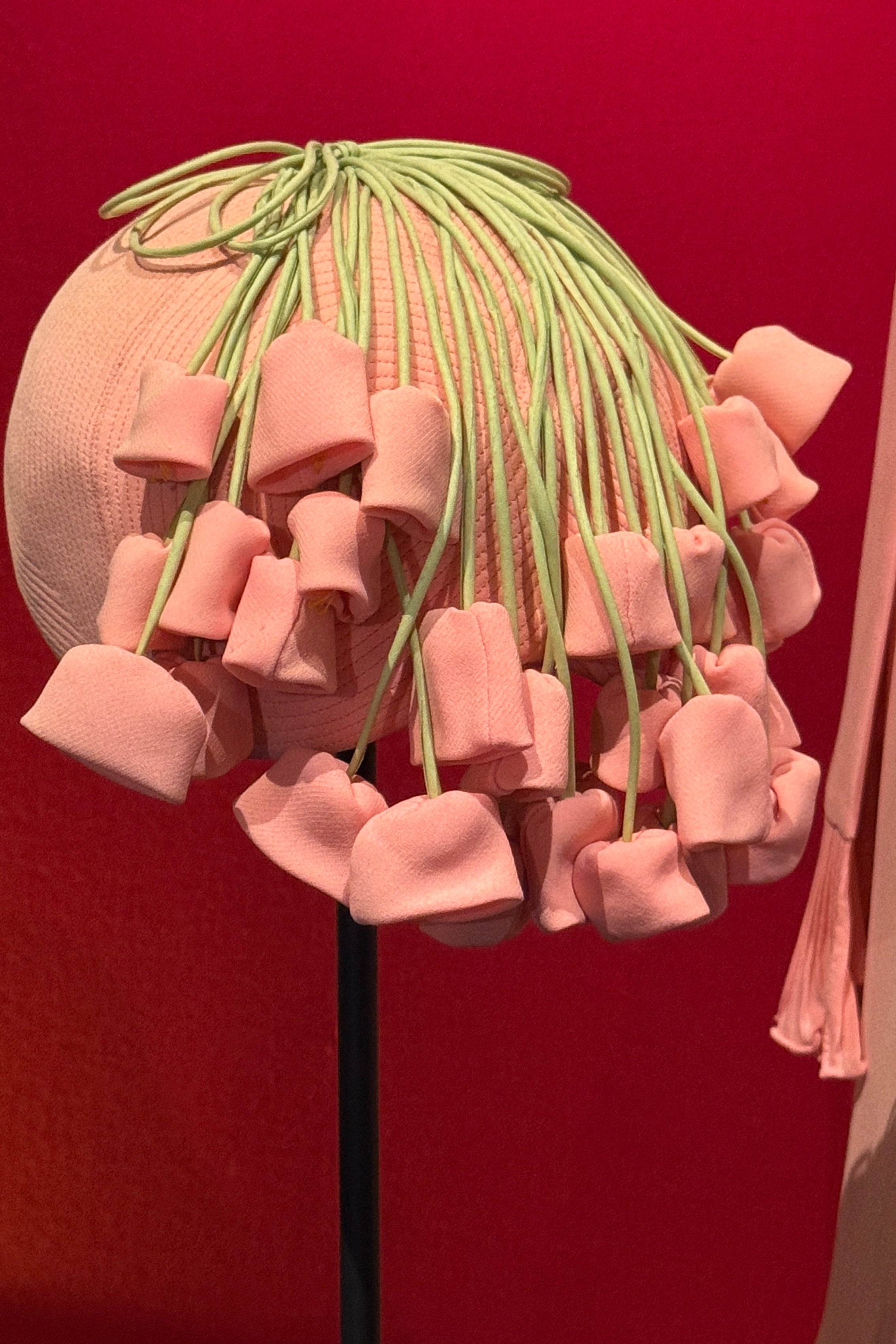 A pink sculptural art piece featuring multiple soft pink flower-like forms hanging from thin green stems, displayed against a solid red background.