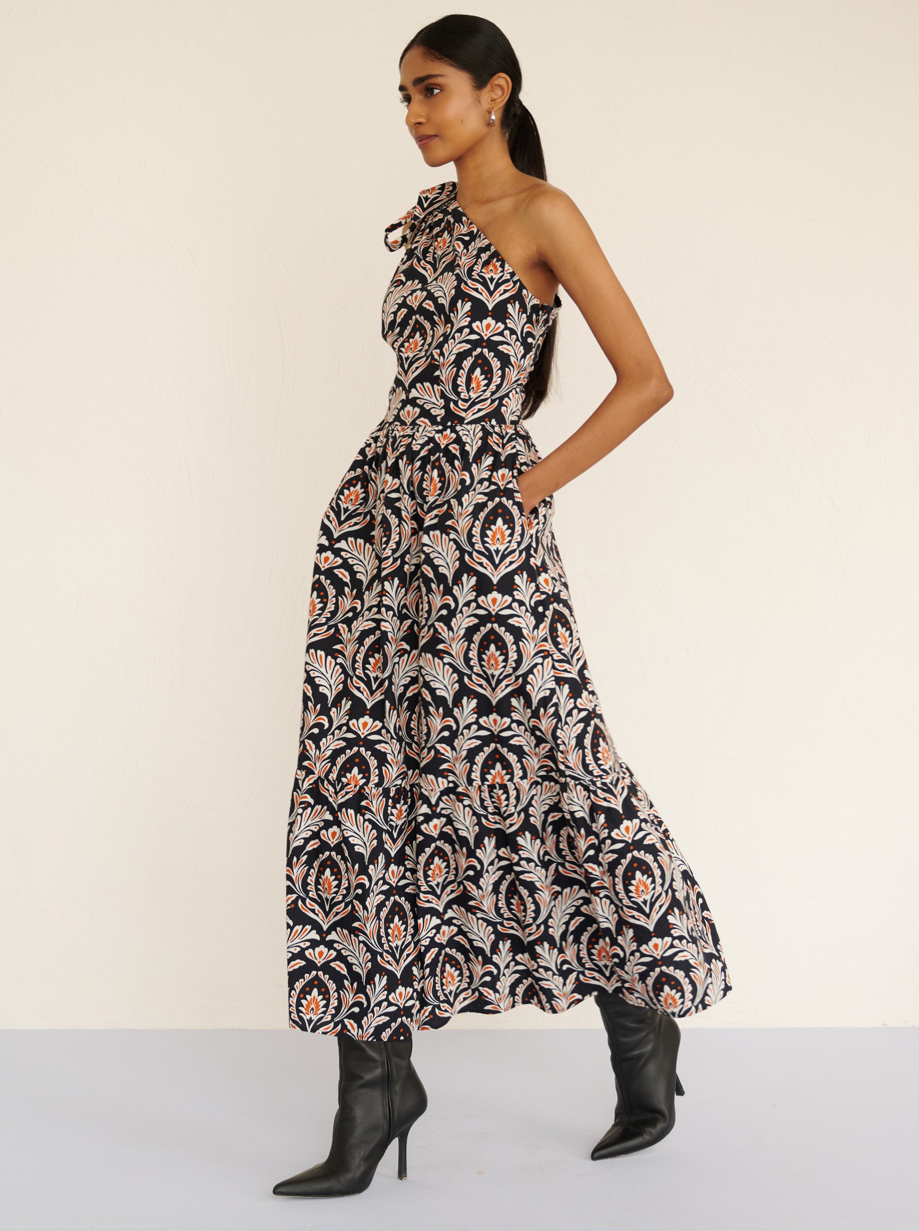 Side view of a model walking. She is wearing the Aliza full-length dress in crisp cotton poplin with the Akbar print. It is featuring an adjustable tie at the shoulder and side-seam pockets.