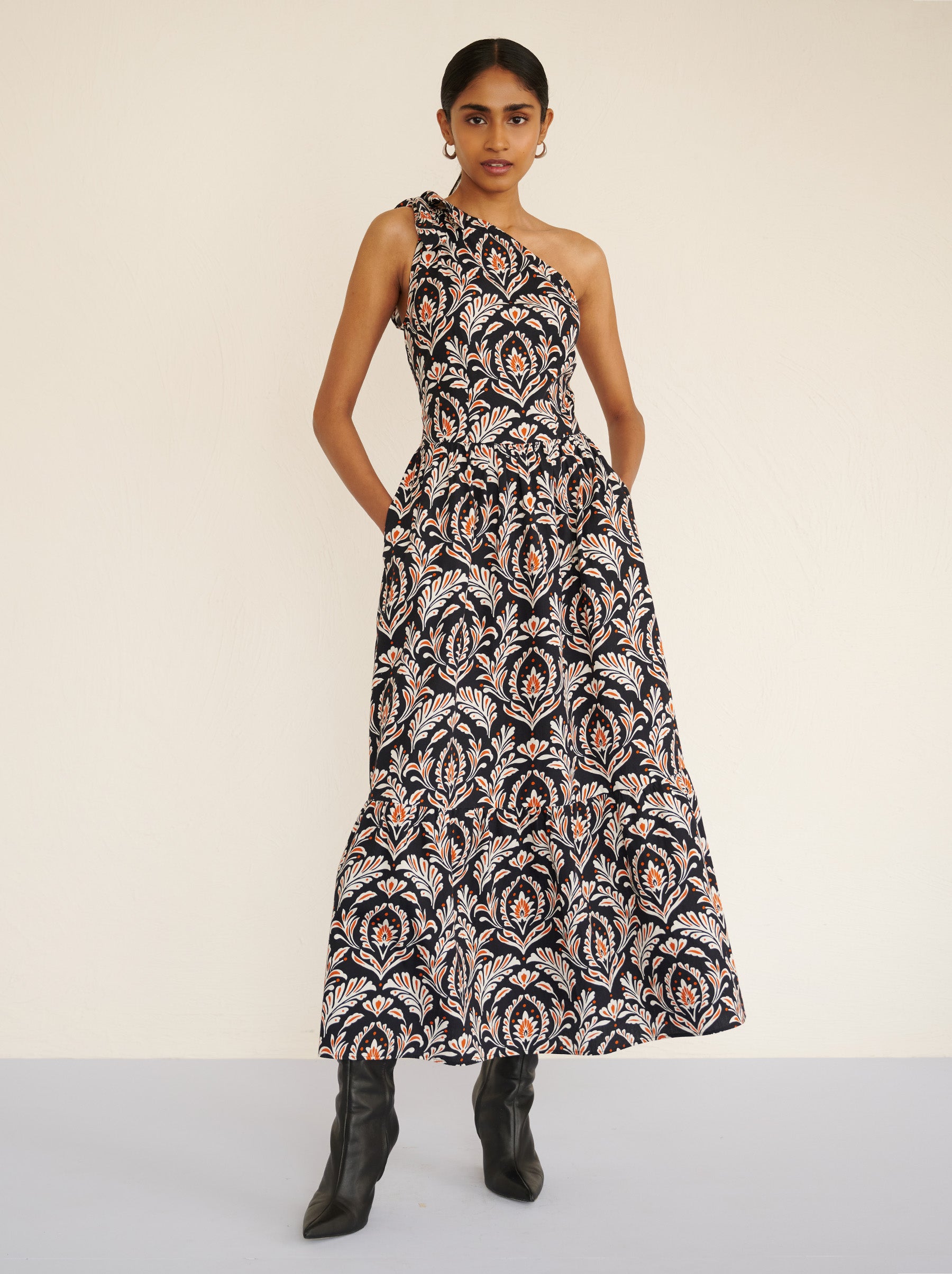 Front view of a model standing with hands in pockets. She is wearing the Aliza full-length dress in crisp cotton poplin with the Akbar print. It is featuring an adjustable tie at the shoulder and side-seam pockets.