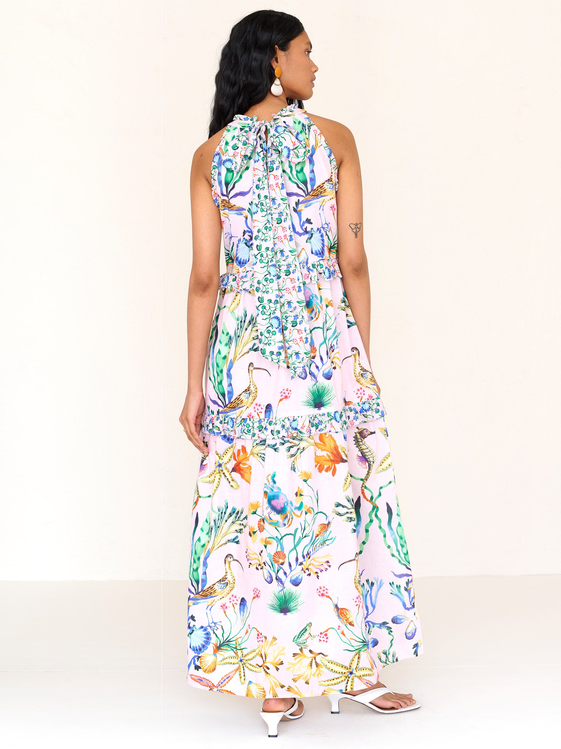Back view of a woman standing. She is wearing Amelia dress in Tidepool print conch color. It features starfish, seahorse, and shell motifs, contrast conch ruffles, and a tie-neck halter back.