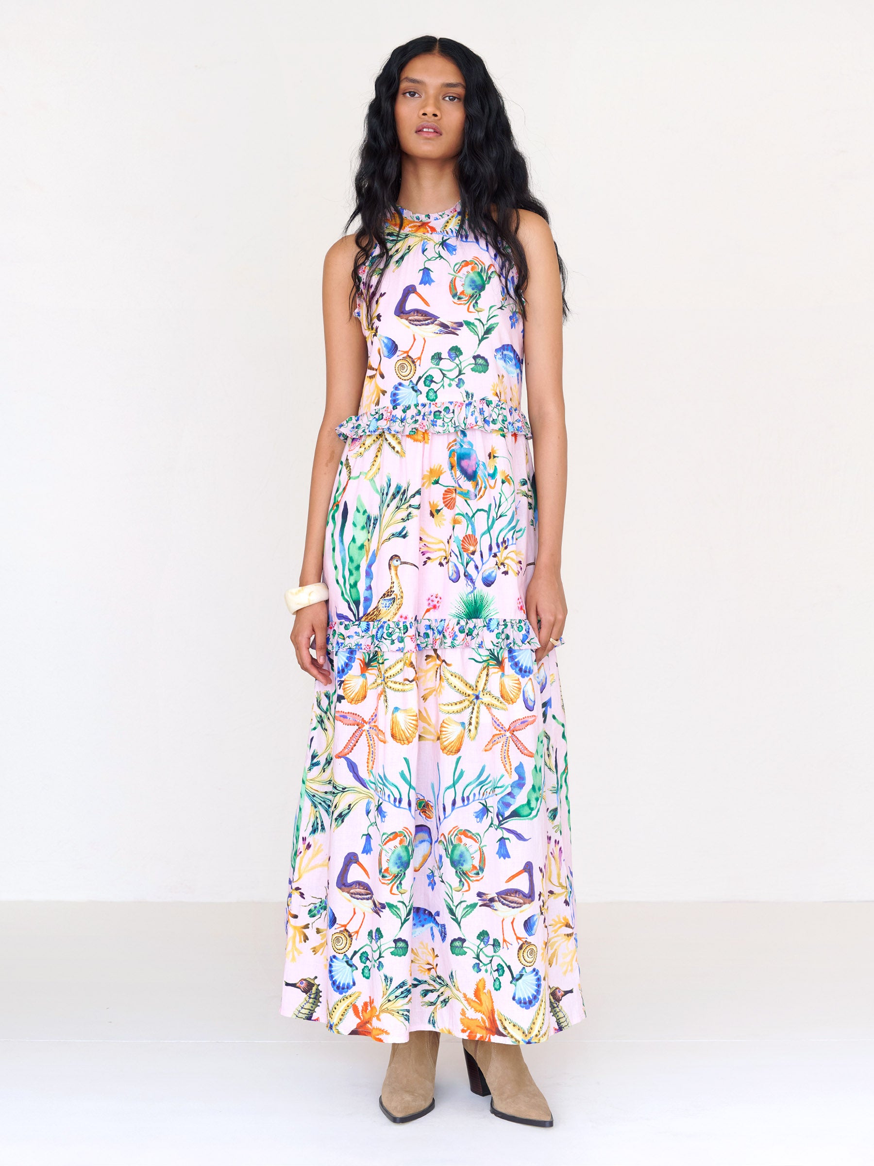 Front view of a woman standing. She is wearing Amelia dress in Tidepool print conch color. It features starfish, seahorse, and shell motifs, contrast conch ruffles, and a tie-neck halter back.