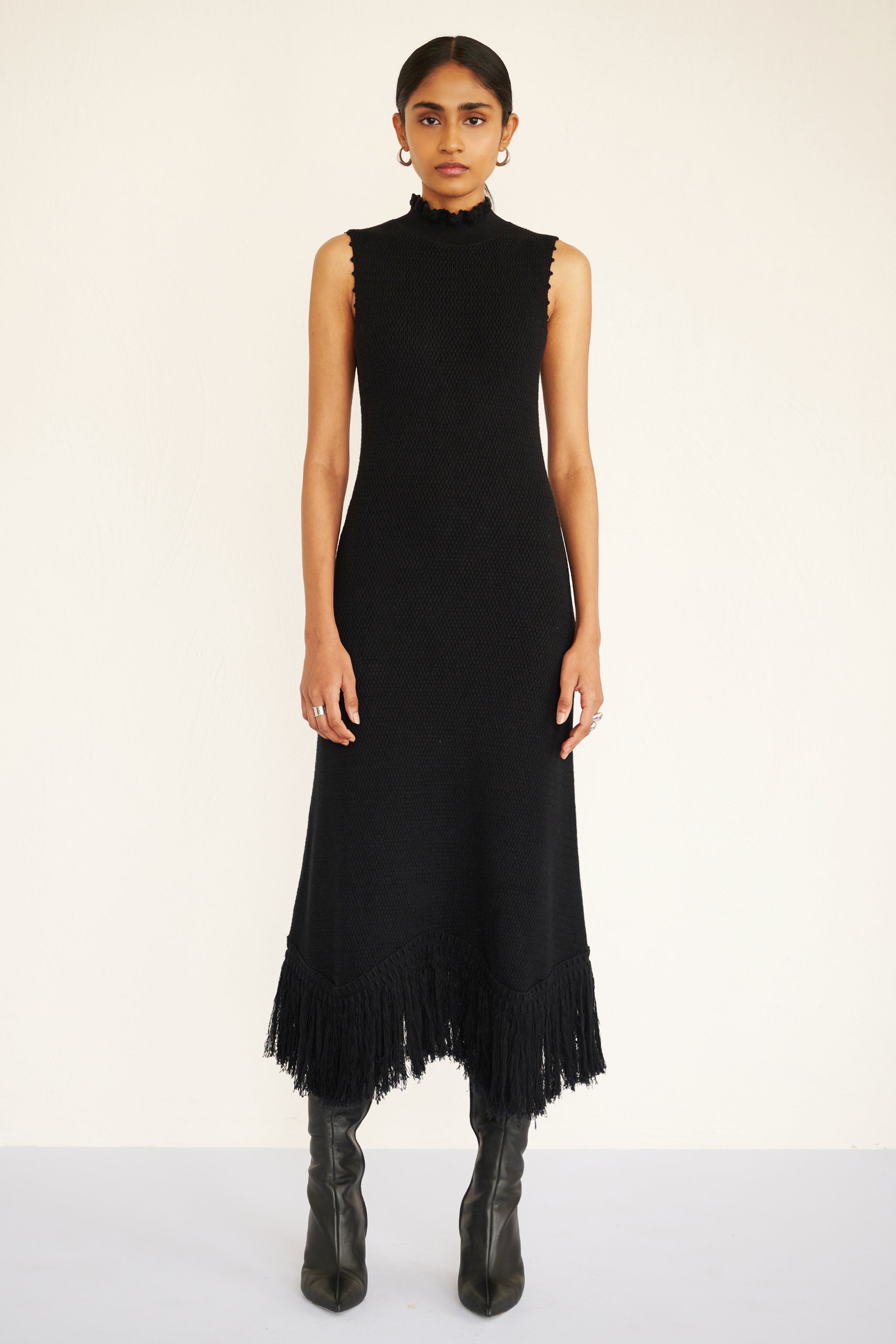 Front view of woman wearing a sleeveless cotton midi dress with a high neckline, hand-crocheted trim, and fringed asymmetrical hem.