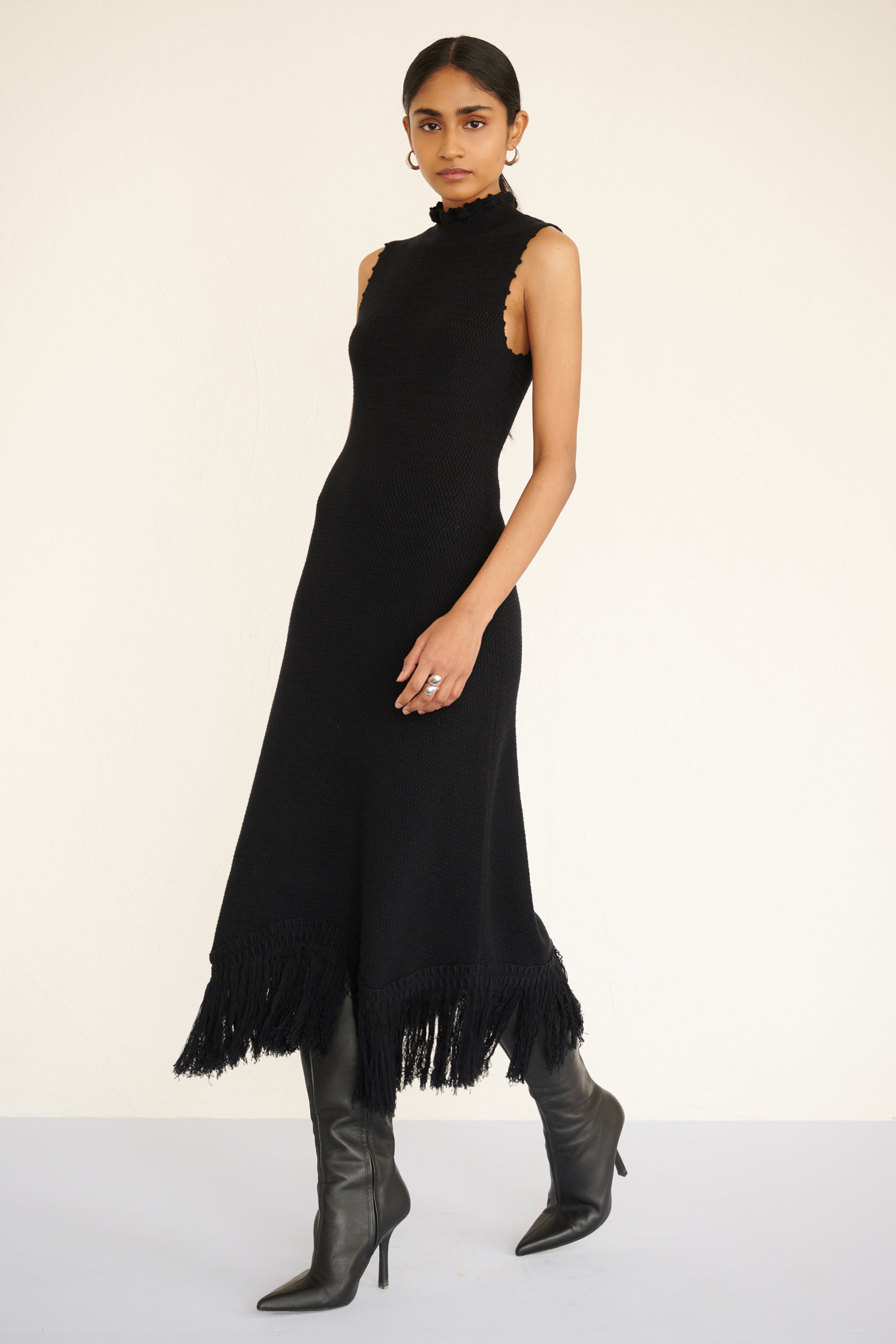Front side view of woman wearing a sleeveless cotton midi dress with a high neckline, hand-crocheted trim, and fringed asymmetrical hem.
