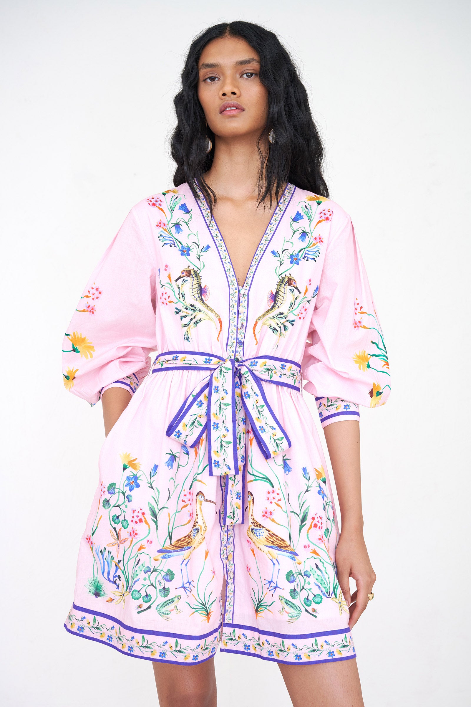 Front view close up of a woman standing wearing the Anne dress in Tidepool print. It features long sleeves, elasticated waist, fabric-covered buttons with loops, fabric belt, side-seam pockets, and a short hemline.