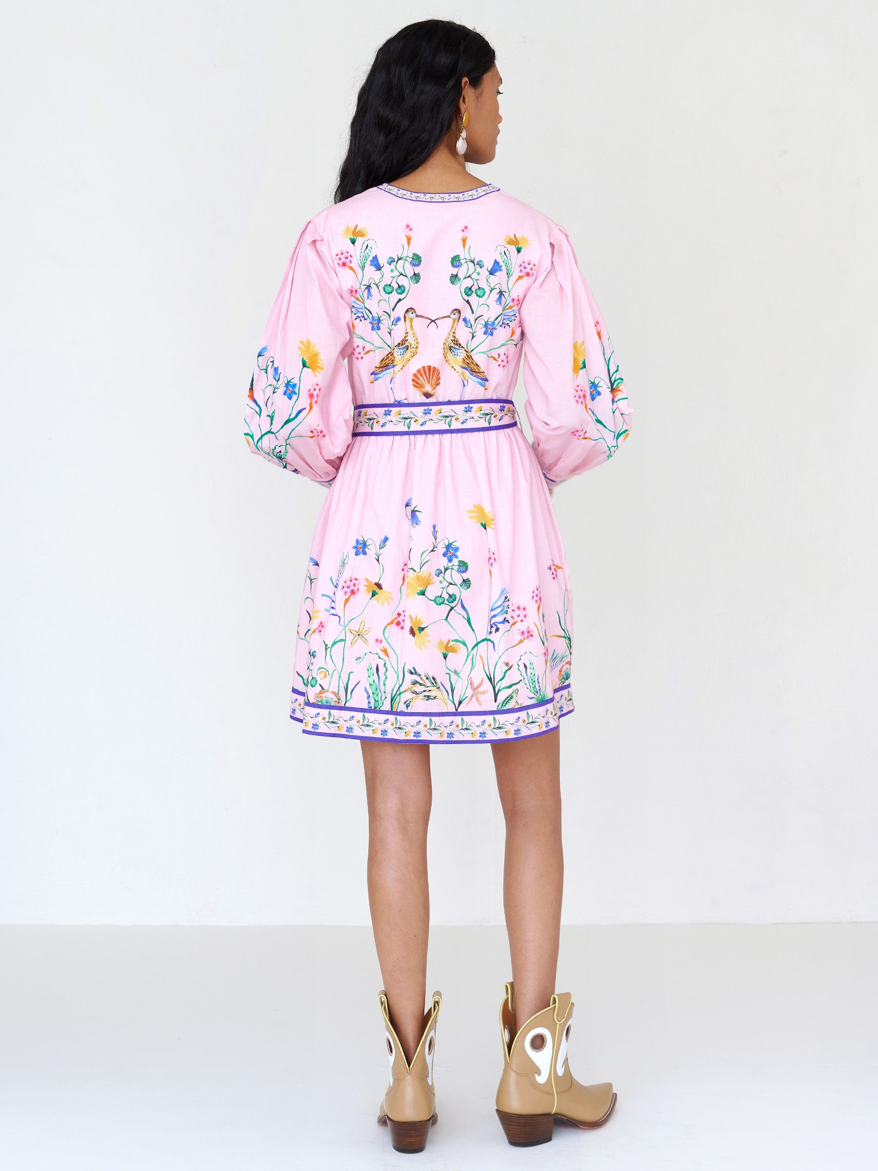 Back view of a woman standing. She is wearing the Anne mini dress in Tidepool print conch color. It features long sleeves, a short hemline, an elasticated waist, fabric-covered buttons, and a fabric belt.